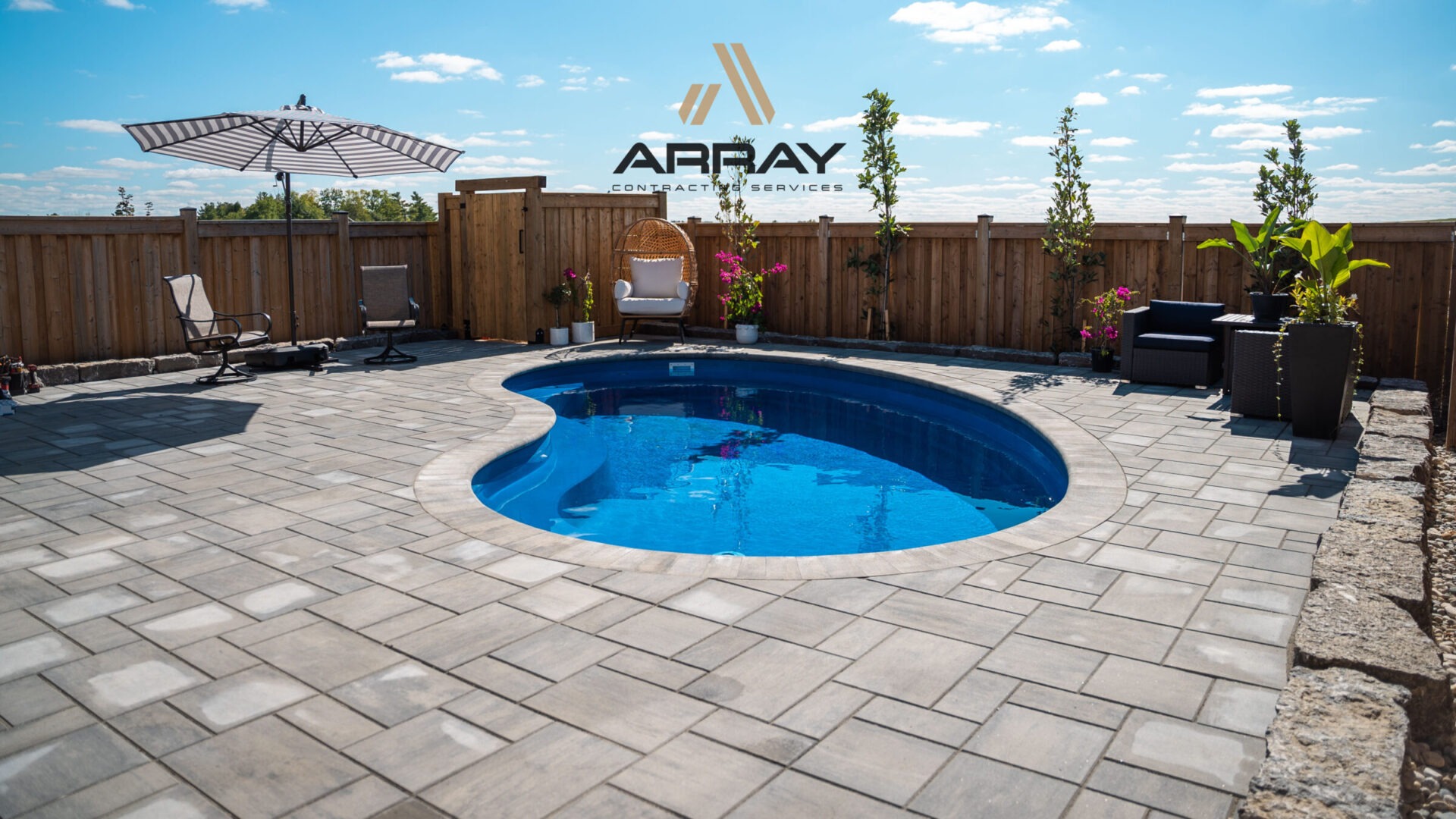 A small backyard pool with stone patio, surrounded by chairs, plants, and wooden fencing under a clear blue sky.