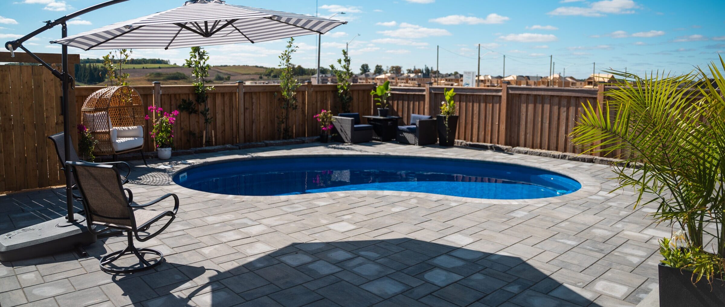 A spacious backyard features a kidney-shaped pool, patio chairs, a large umbrella, potted plants, and wooden fencing against a clear blue sky backdrop.