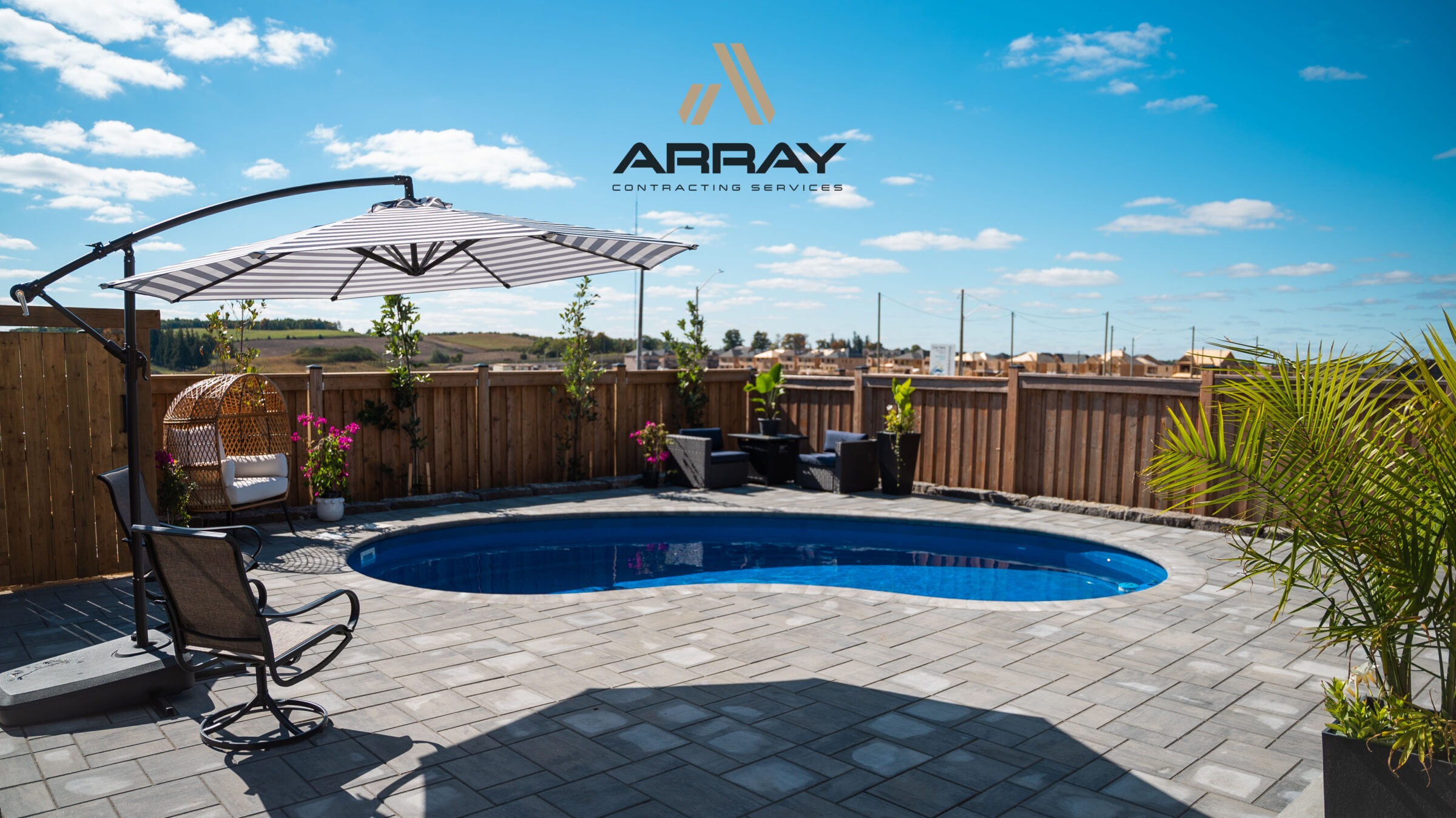 A backyard pool with patio furniture, umbrella, and plants, enclosed by a wooden fence under a clear blue sky. No landmarks visible.