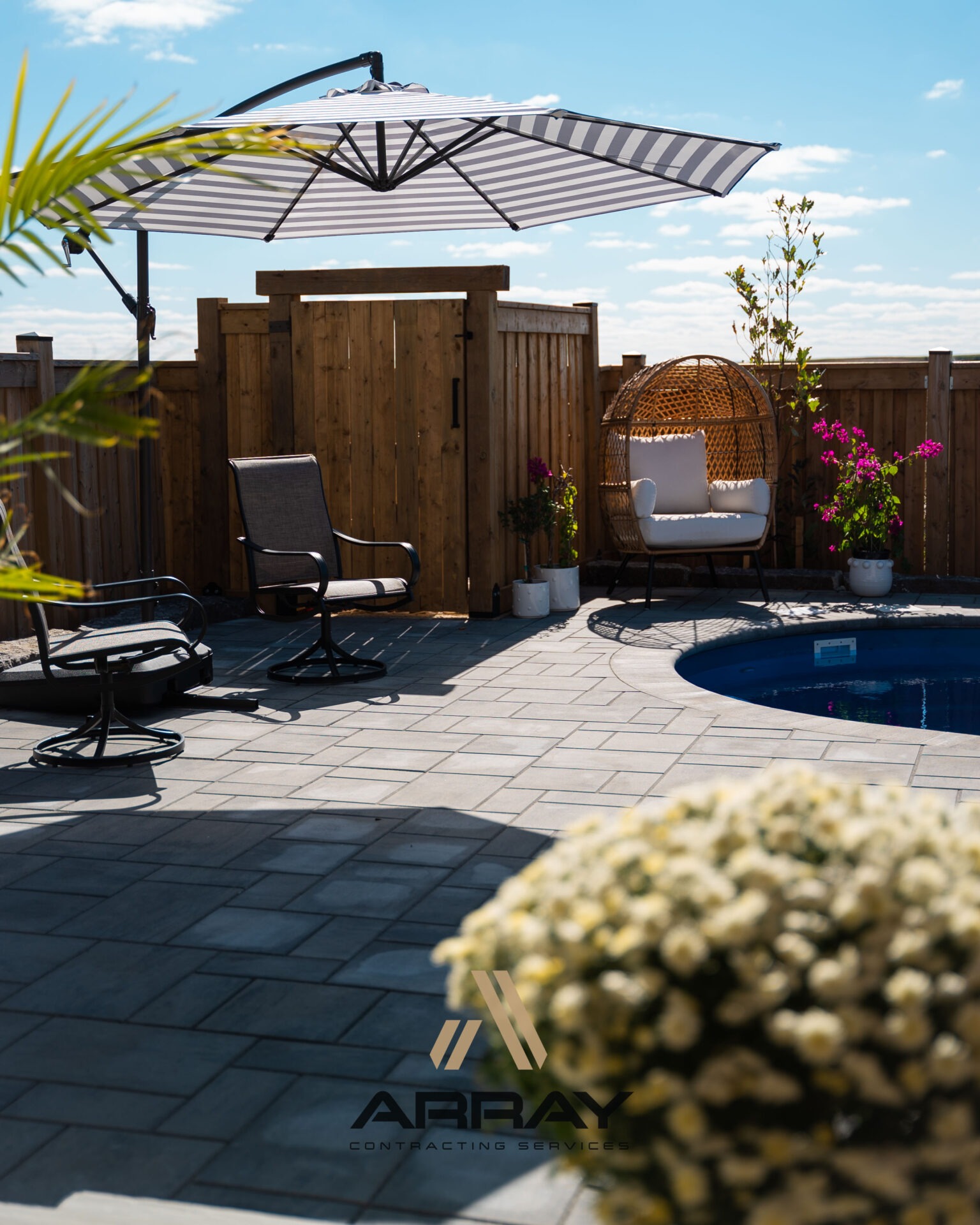 A backyard scene with a pool, lounge chairs, and a striped umbrella, surrounded by a wooden fence and potted plants.
