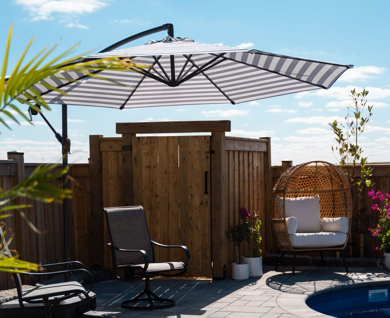 A sunny patio features a striped umbrella, comfortable chairs, and potted plants, creating a relaxing atmosphere beside a circular pool.