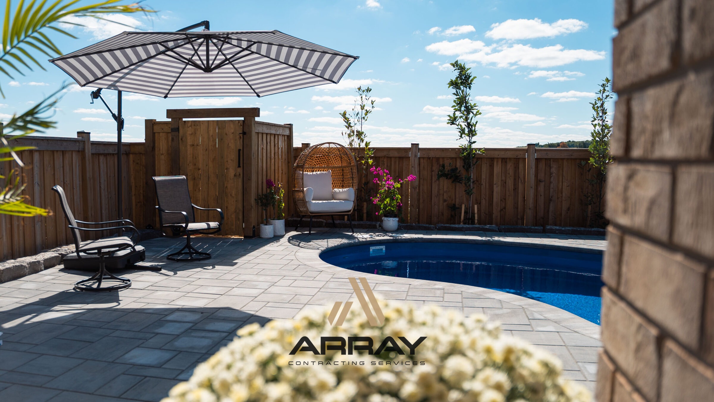Backyard with a small pool, patio chairs, umbrella, and plants. Wooden fence surrounds the area. Clear blue sky overhead. No people present.