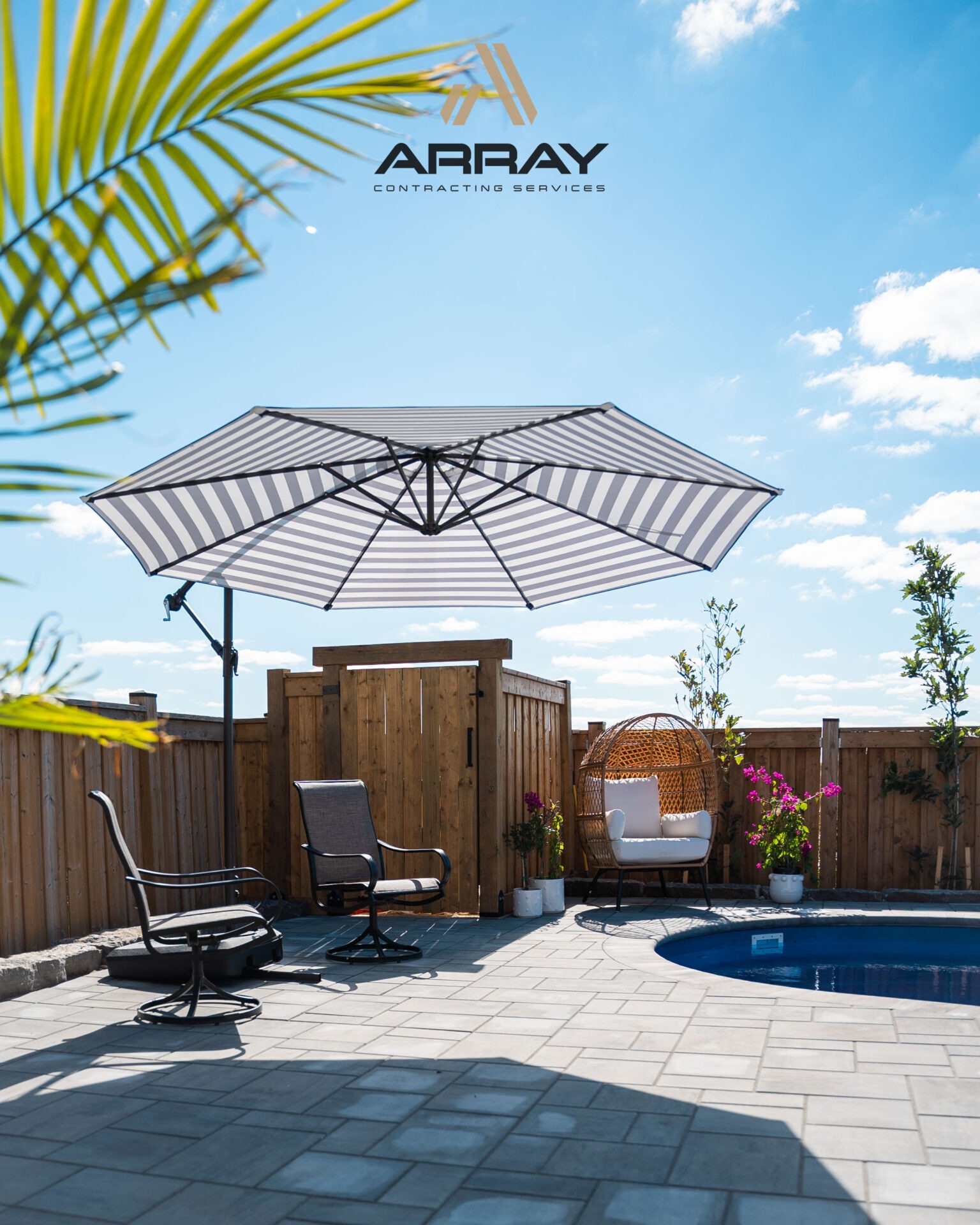 Outdoor patio with striped umbrella, seating, and potted plants by a pool. Clear blue sky in the background, no recognizable landmarks visible.
