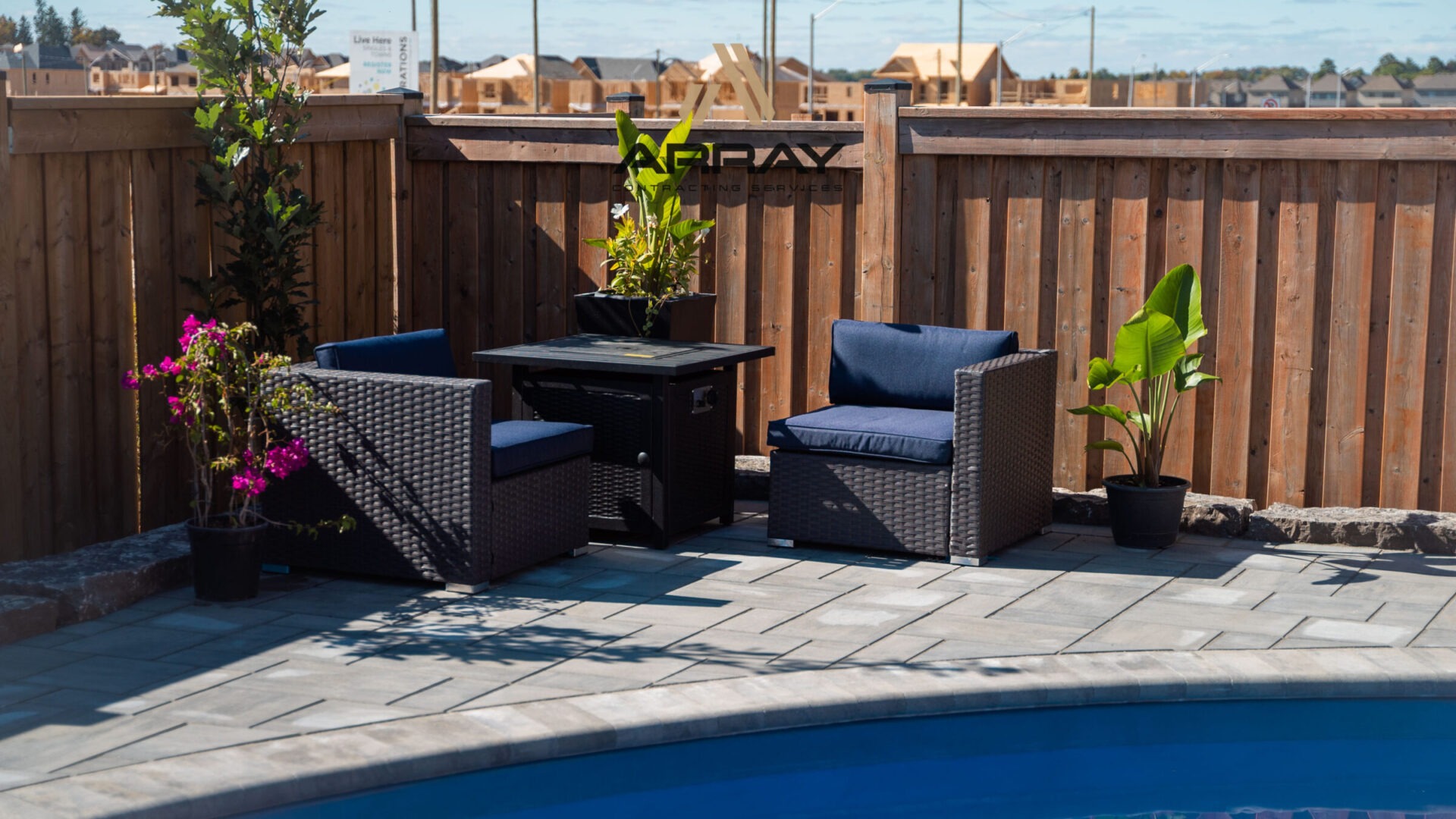 Outdoor patio with two chairs, small table, and potted plants beside a pool, enclosed by a wooden fence and brick paving.