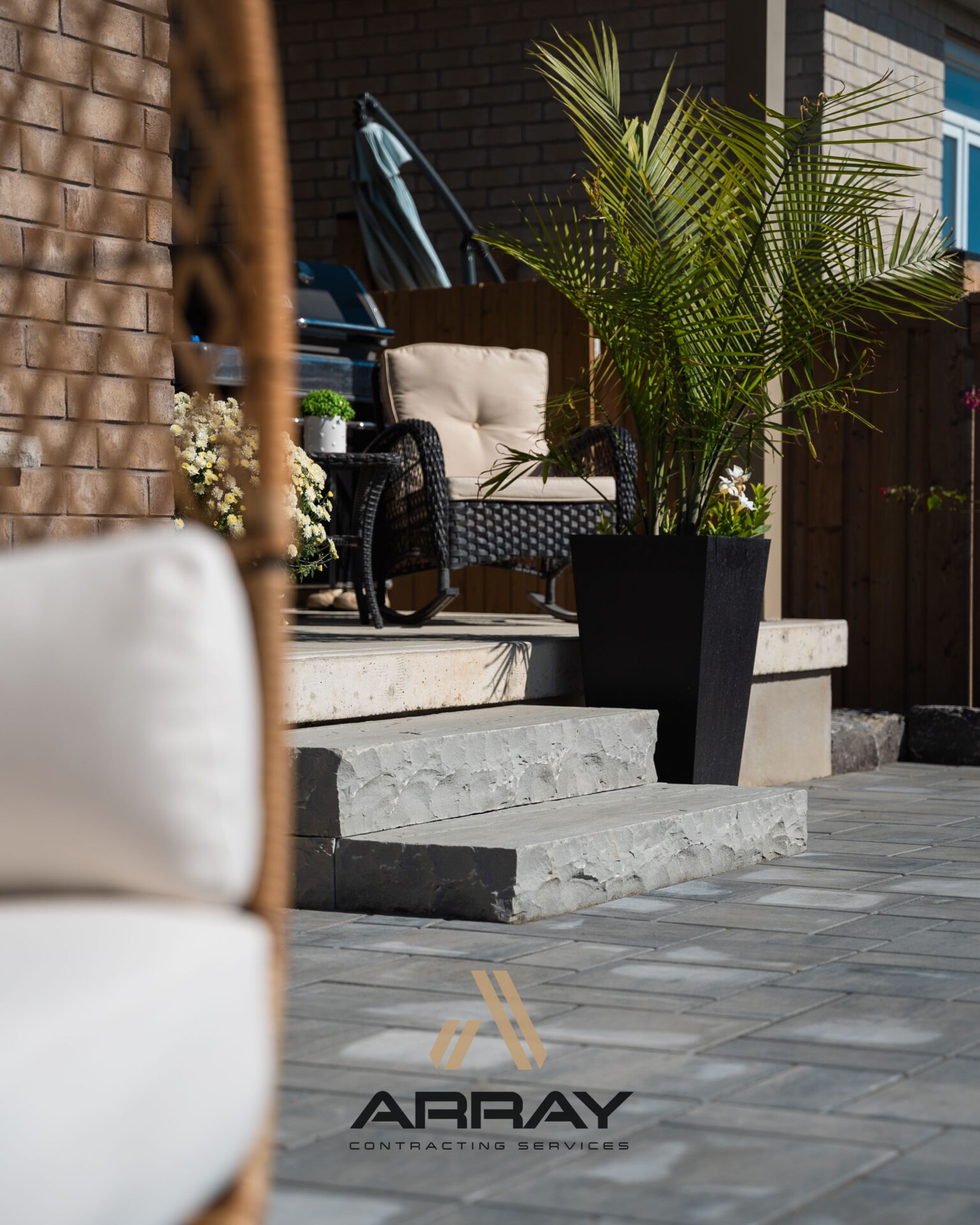 A patio with stone steps, wicker furniture, potted plants, and a decorative logo. The setting is sunny with modern design elements.