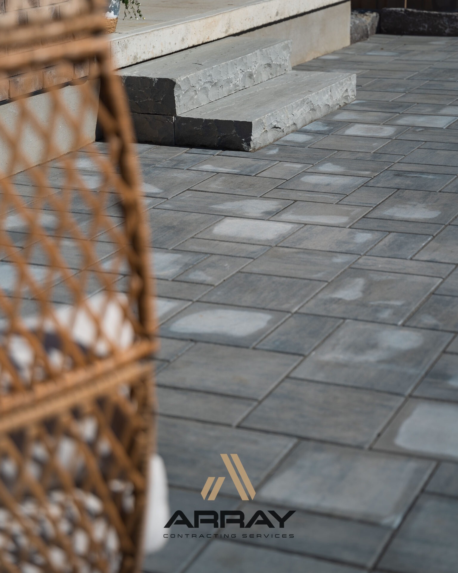 Modern patio with stone tiles and steps, featuring a wicker chair in the foreground. Logo: 
