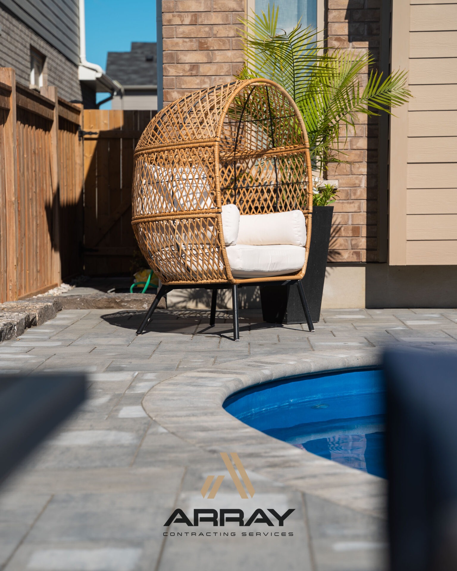 Cozy patio scene with a rattan chair, potted plant, and small swimming pool. Sunlit stone tiles enhance the modern outdoor setting with privacy fencing.