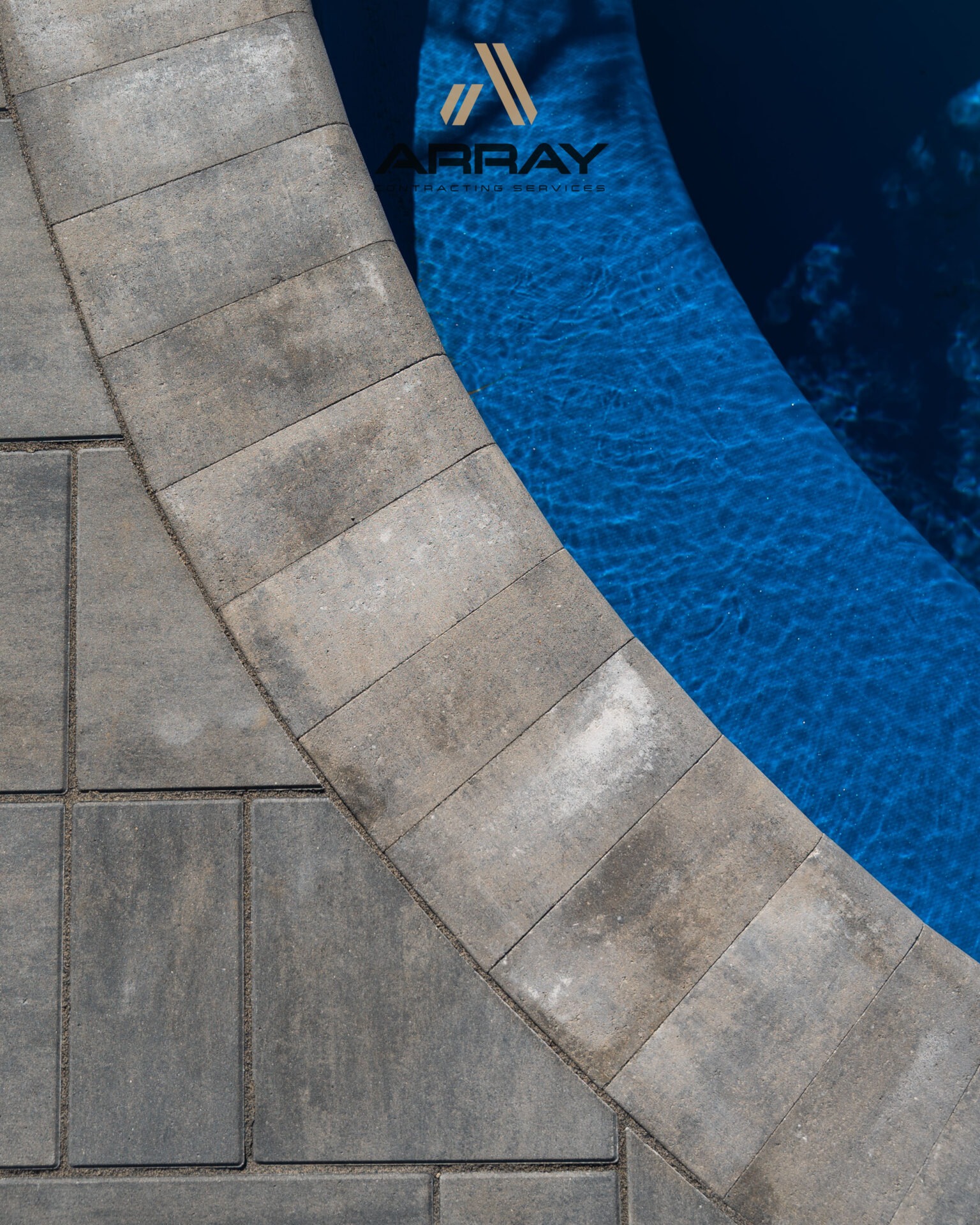 A curved stone pathway surrounds a clear blue pool. The surface features neat arrangements of rectangular tiles and a company logo.