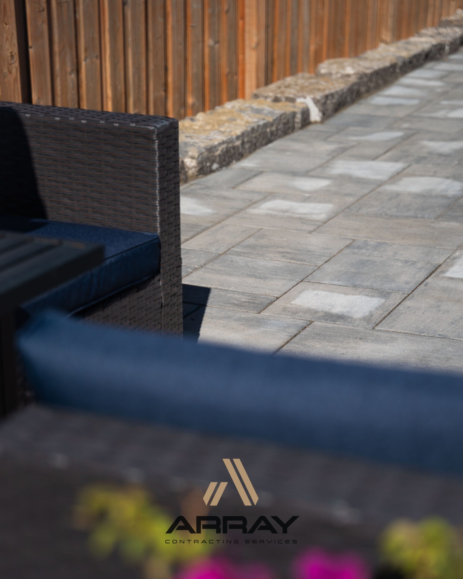 A neatly arranged patio features wicker furniture, gray paving stones, and a wooden fence. The branding 