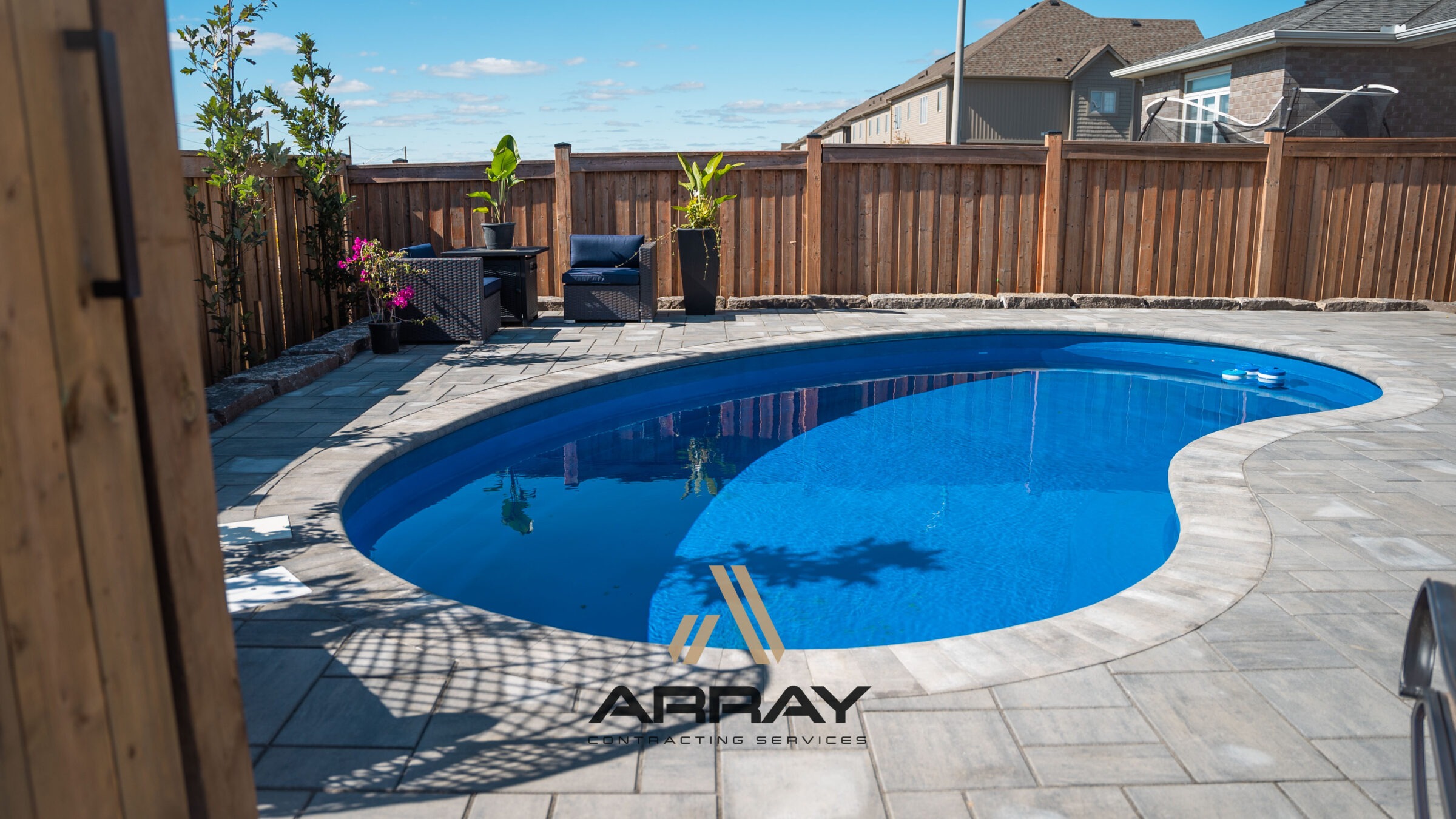 A backyard features a blue kidney-shaped pool, surrounded by stone tiles, potted plants, and comfortable seating next to a wooden fence.
