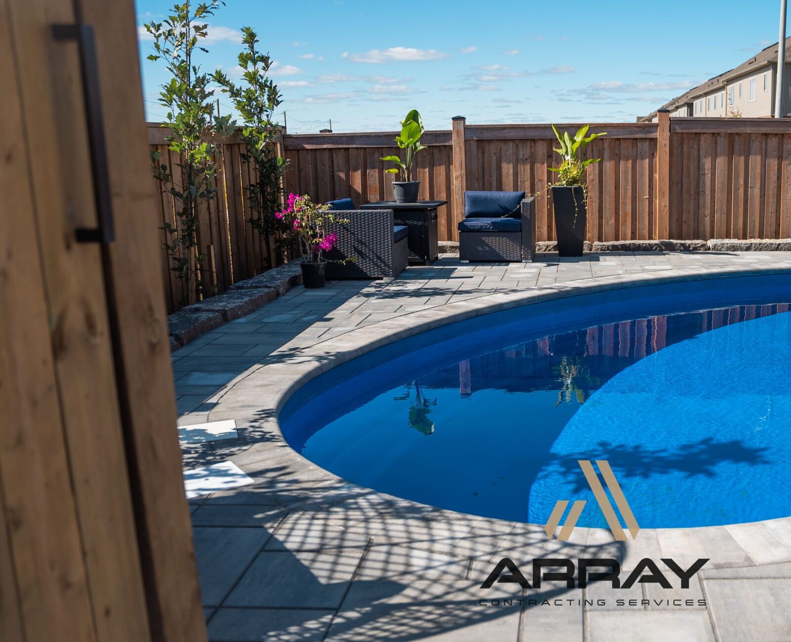 A modern backyard features a kidney-shaped pool, patio seating, and potted plants, with wooden fencing and neighboring houses in the background.