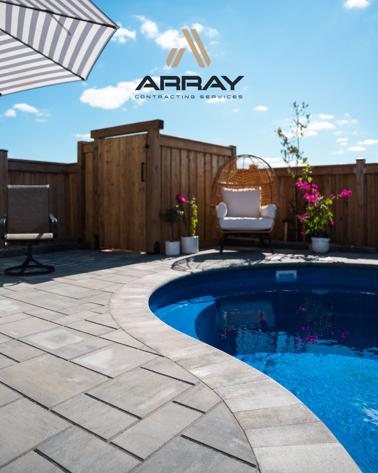 A sunny backyard with a pool, paver patio, striped umbrella, and wicker chair, surrounded by wooden fencing and vibrant pink flowers.