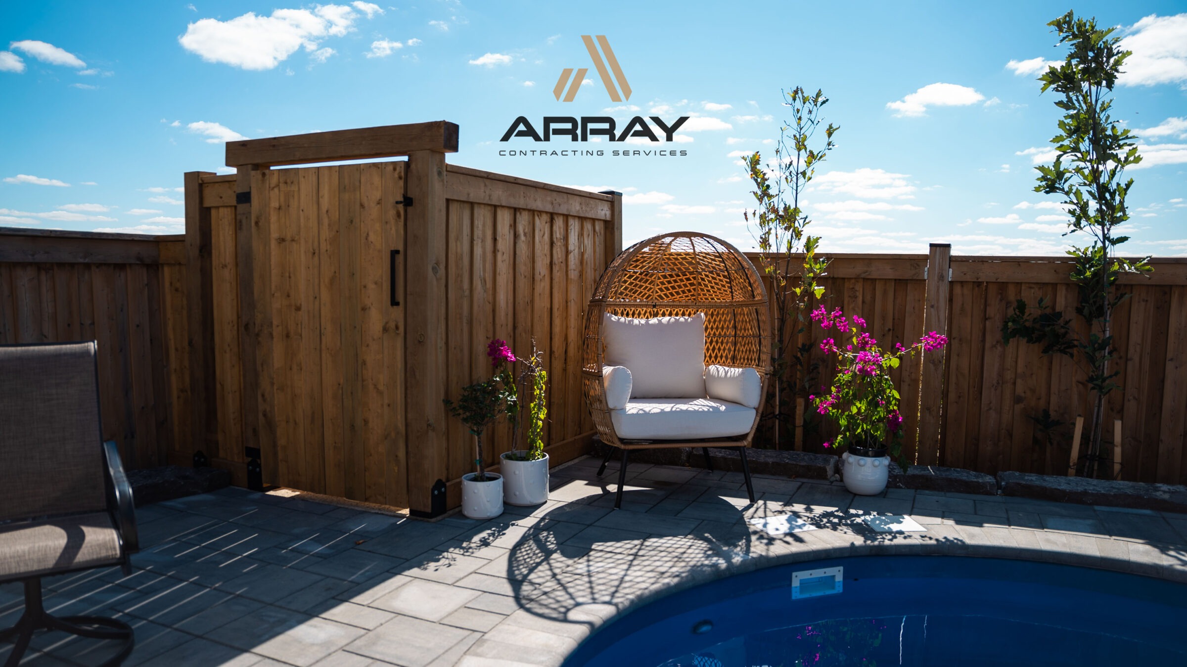 Cozy patio scene with wicker chair, vibrant flowers, and a small pool, set against a wooden fence under a clear blue sky.