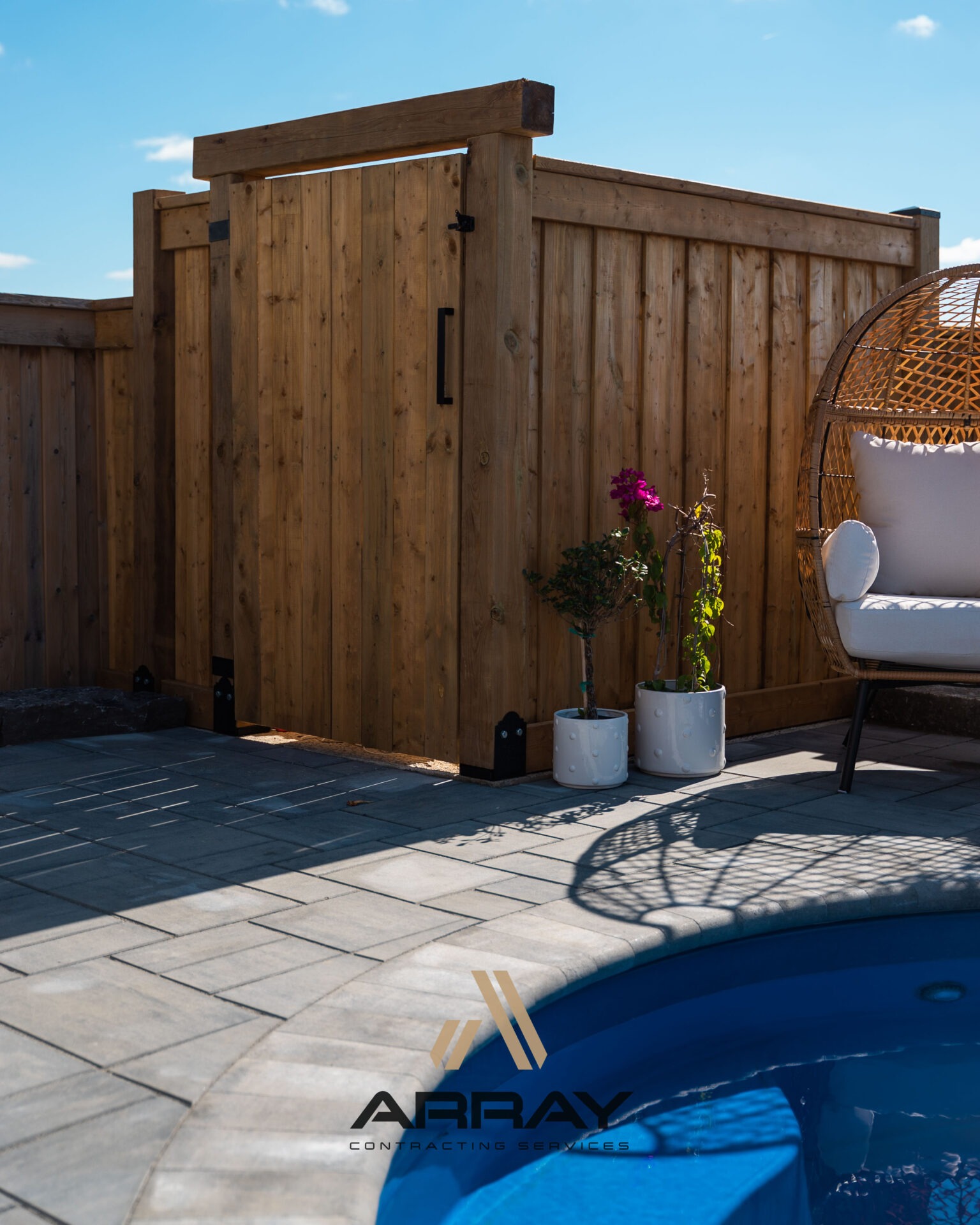 A modern poolside setting features wooden fencing, a hanging chair, potted plants, and elegant paving under a bright blue sky.