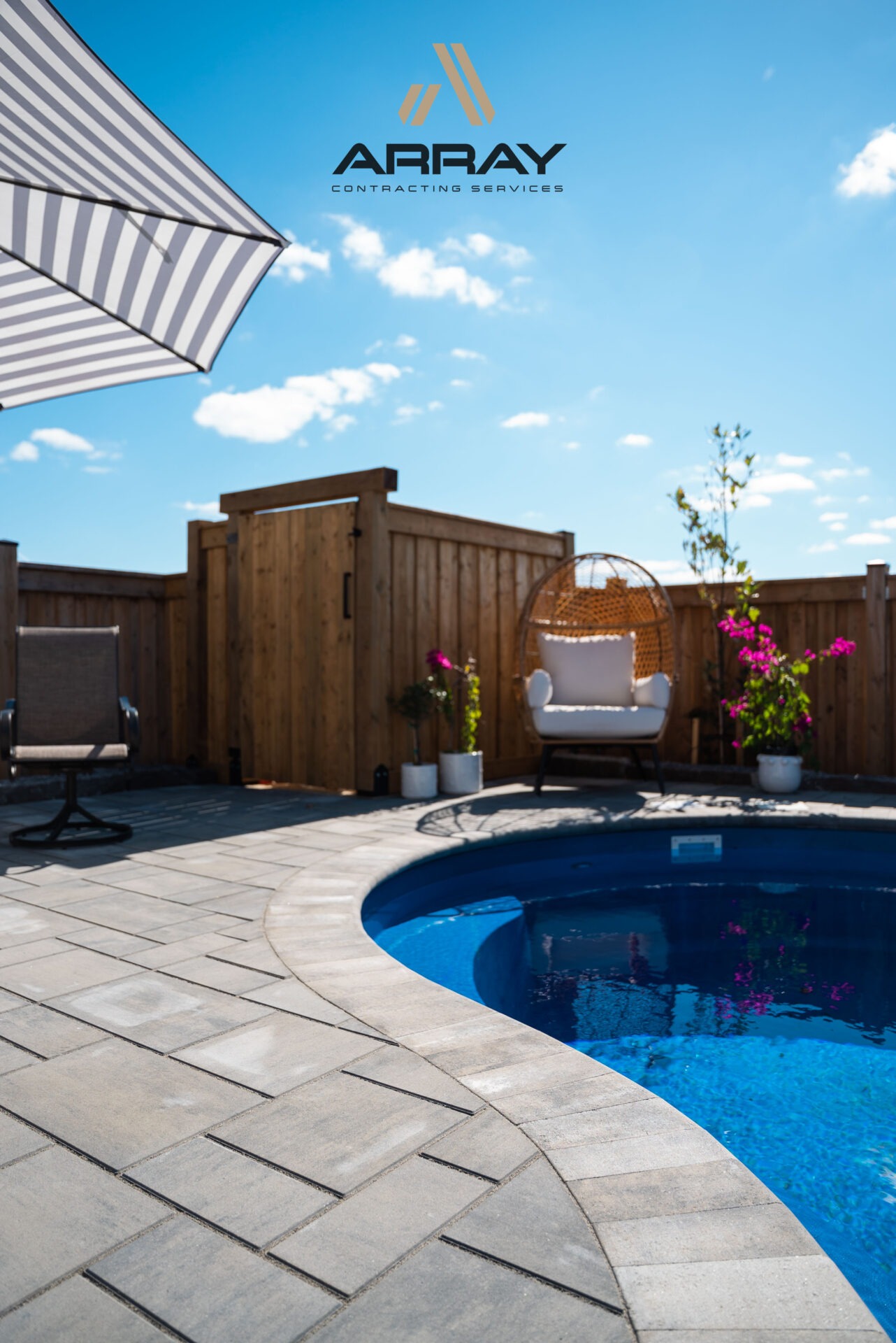 A sunny backyard with a private pool, wooden fence, lounge chairs, and an umbrella, surrounded by potted plants and clear blue sky.
