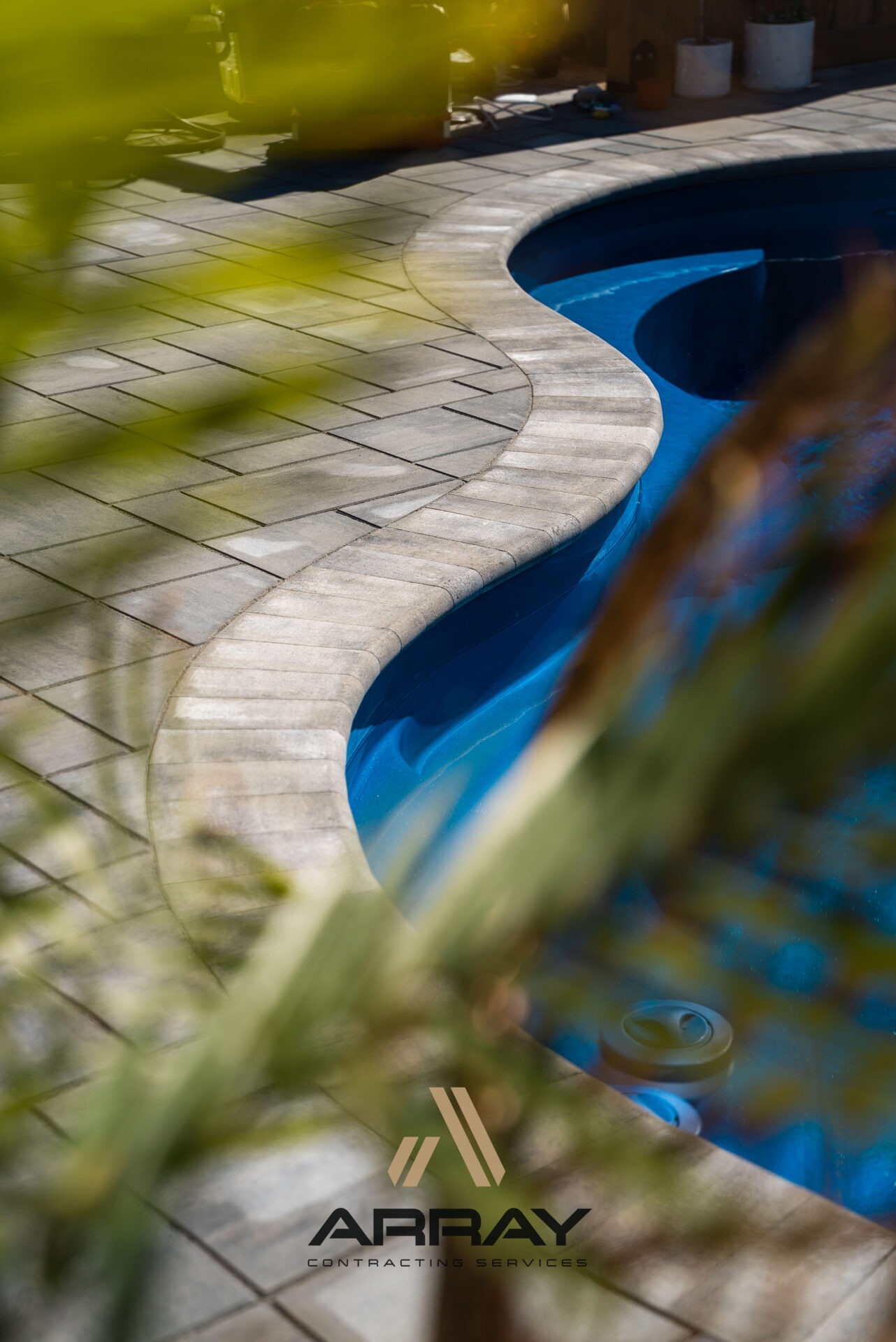 Curved, stone-paved poolside path with blue water through lush green foliage; logo 