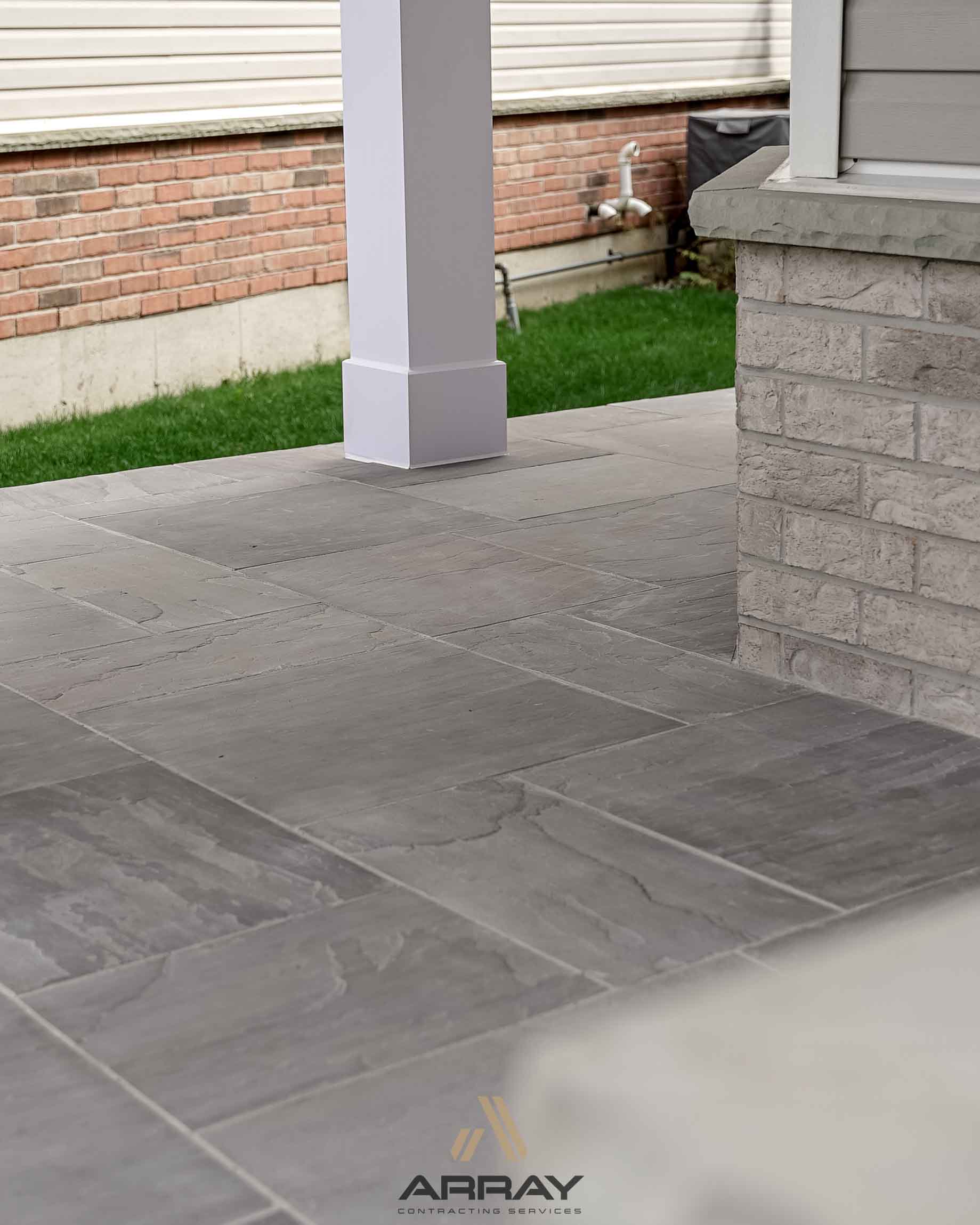 Stone patio featuring gray tiles, white column, and brickwork. Green grass borders the area. Residential setting without recognizable landmarks or historical buildings.