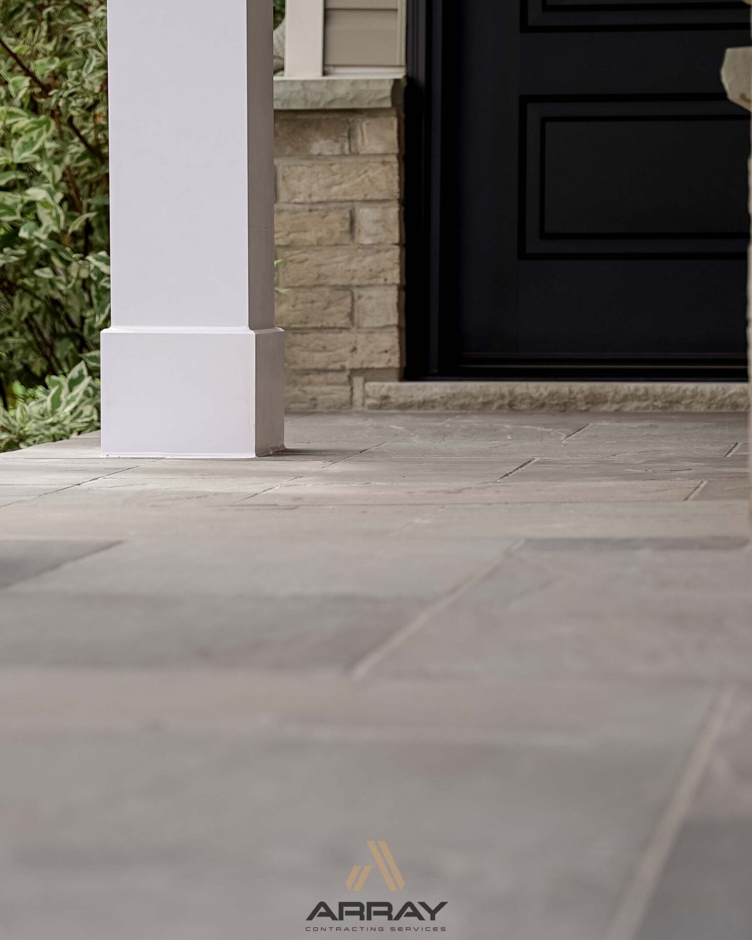 Close-up of a residential porch featuring stone tiles, a white column, stone wall, and a black door. Lush greenery is visible nearby.