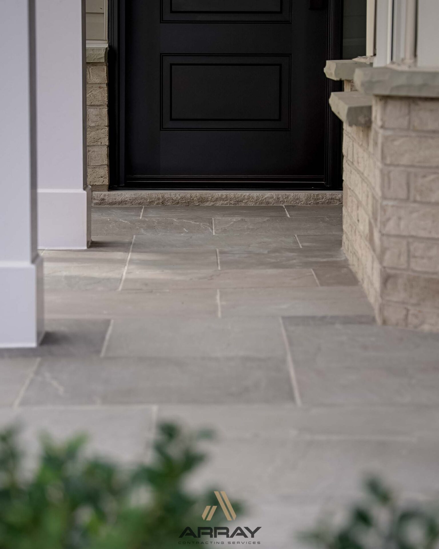 Brick exterior with stone tiles leading to a closed black door. Foreground shows blurred green foliage. Logo reads 