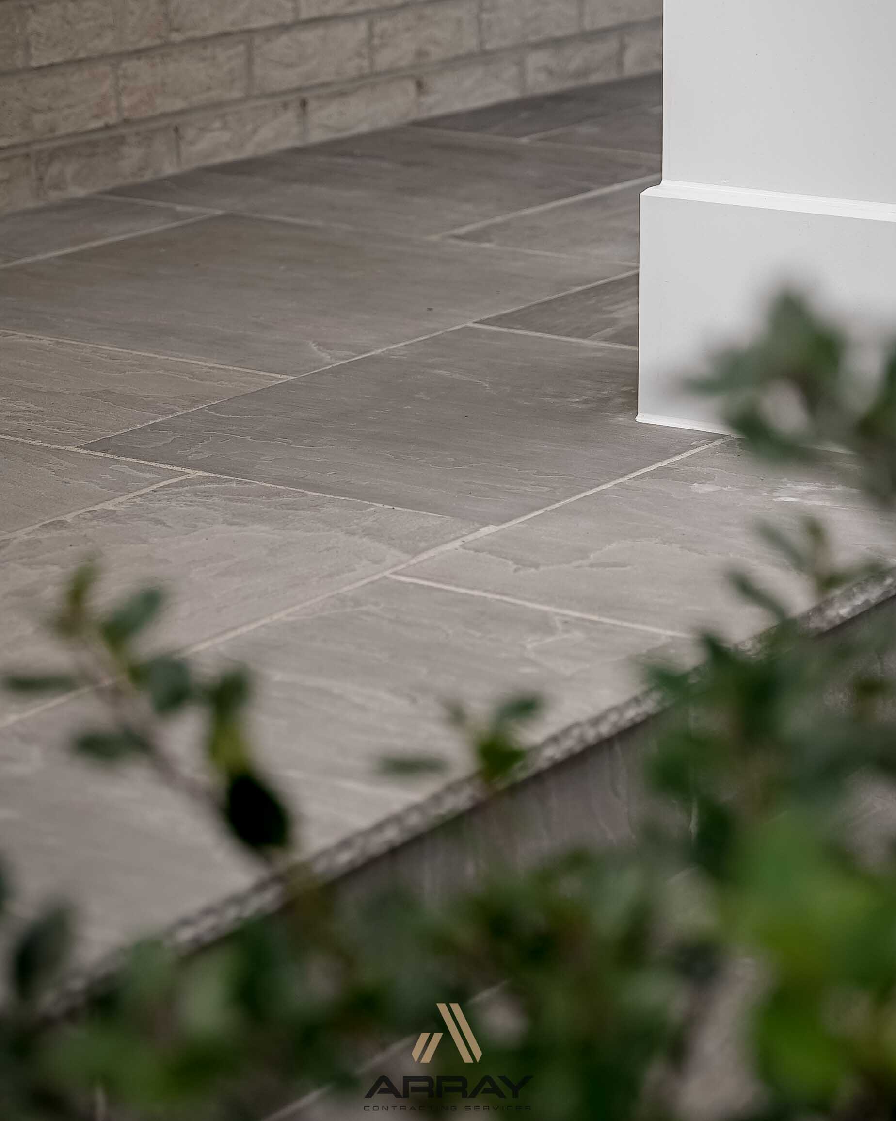 A close-up of stone tile flooring with a white pillar and blurred greenery in the foreground. Cozy and modern architectural elements.