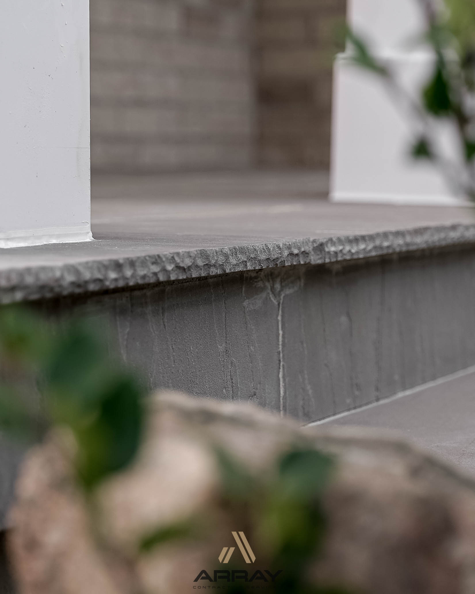 A close-up of stone steps with a rough edge, showing part of a pillar. A blurred plant is in the foreground.