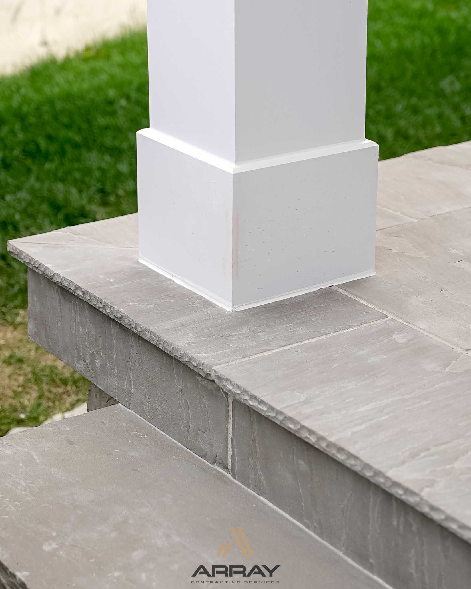 A white column base is supported by textured stone steps, surrounded by a green lawn, representing modern architectural design elements.