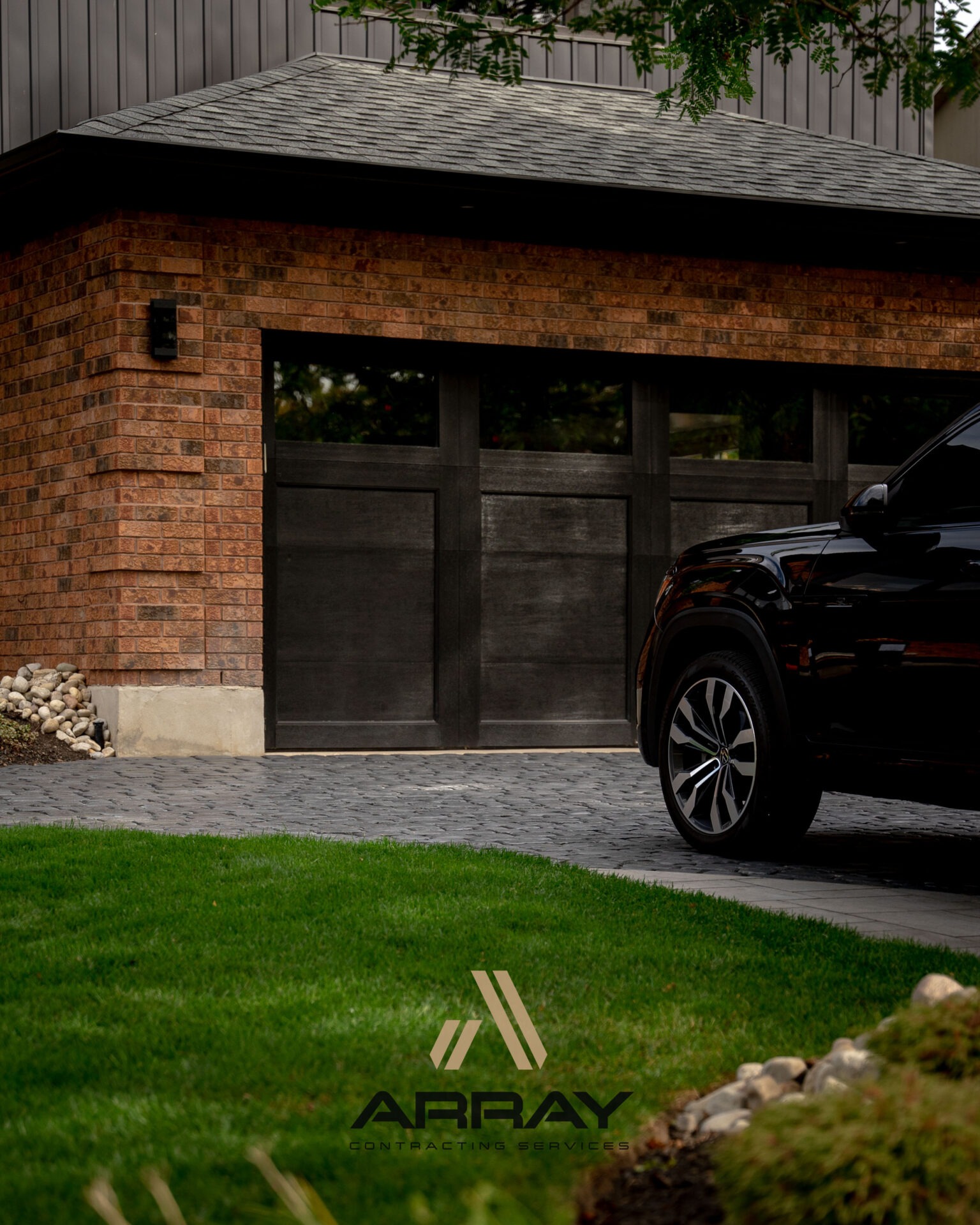 A black vehicle is parked on a brick driveway in front of a brick garage, with a well-maintained grassy lawn nearby.