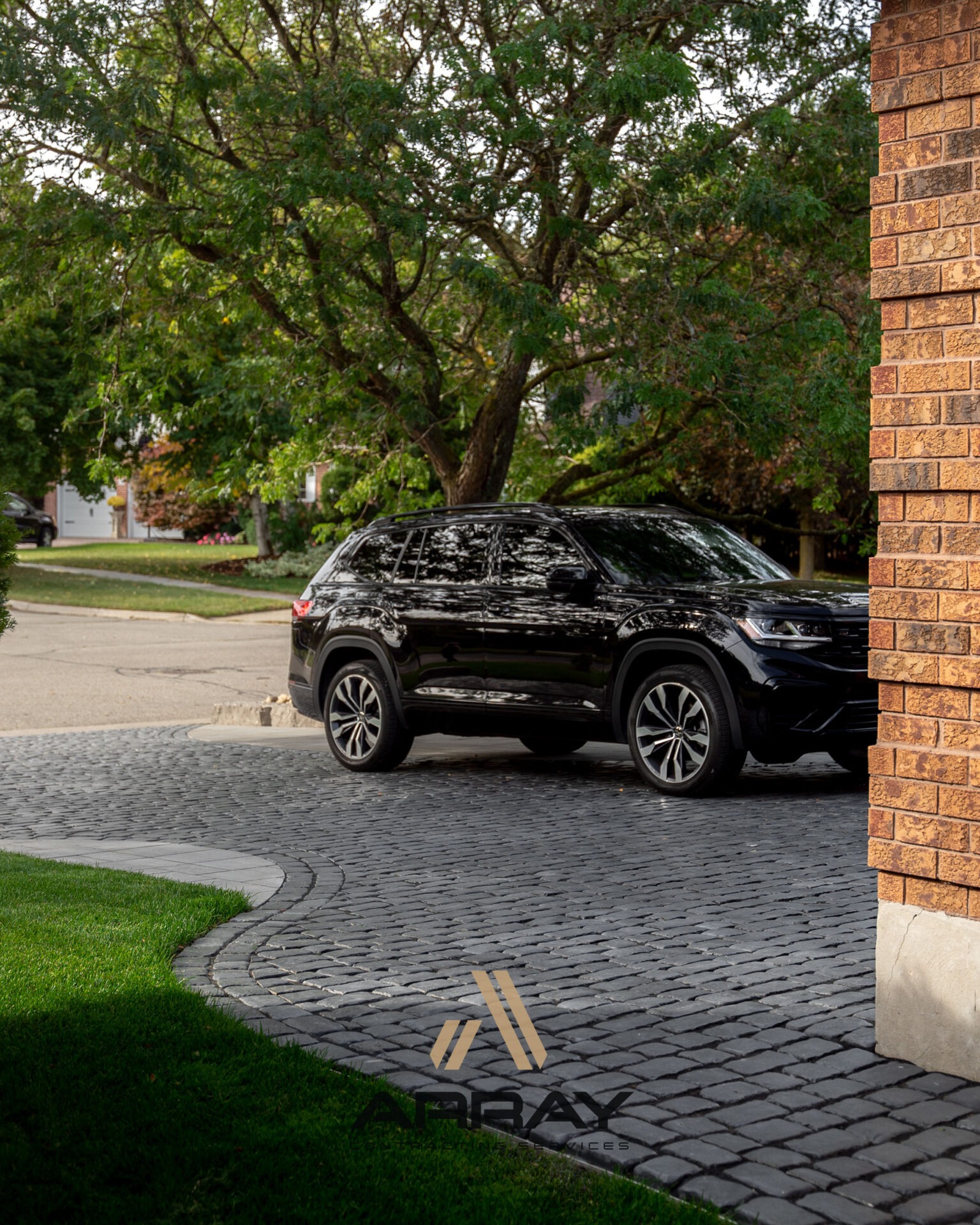 A black SUV parked on a cobblestone driveway beside a brick house, surrounded by lush trees and a grassy lawn.