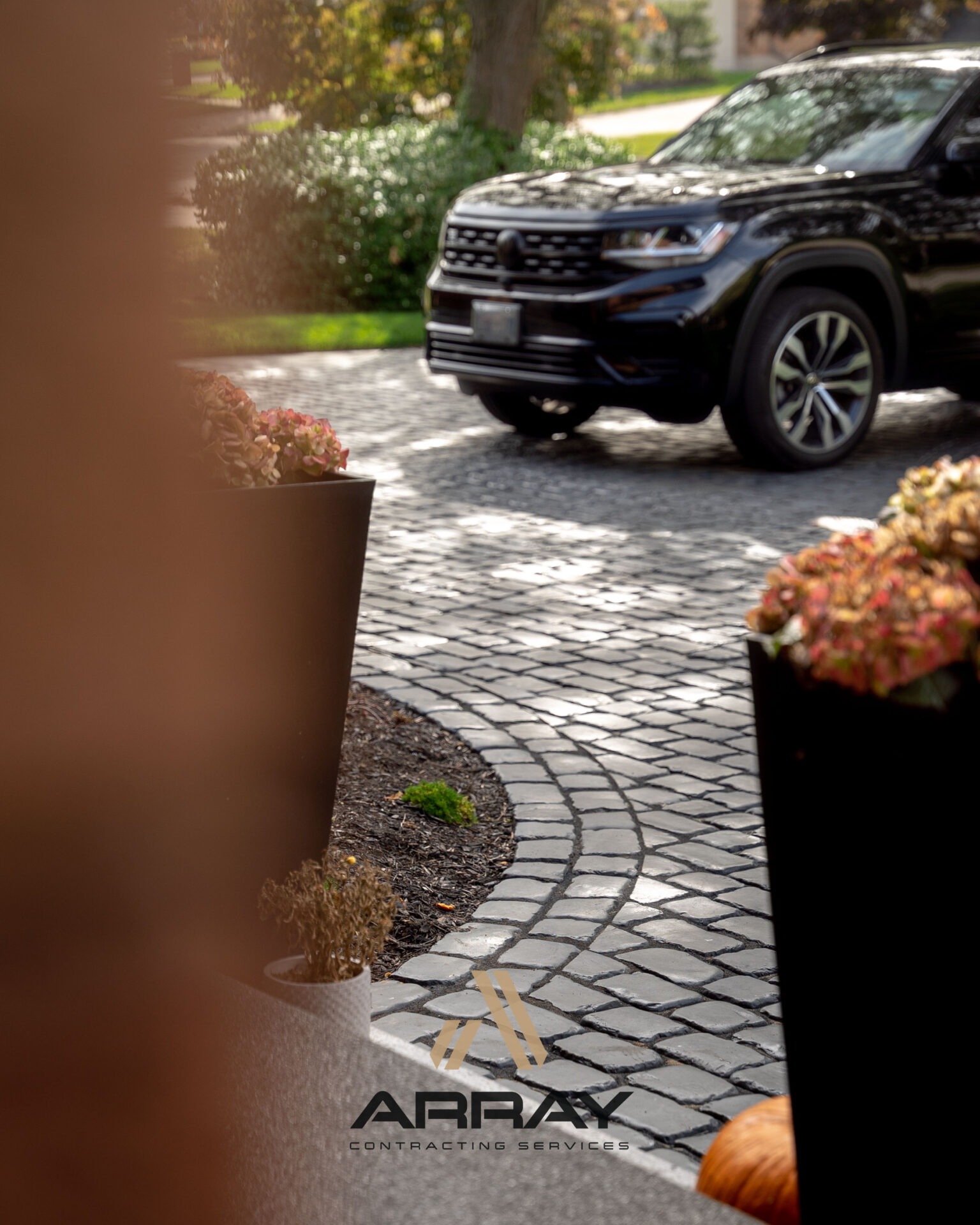 A black SUV parked on a cobblestone driveway, surrounded by potted plants and greenery, with visible company branding 