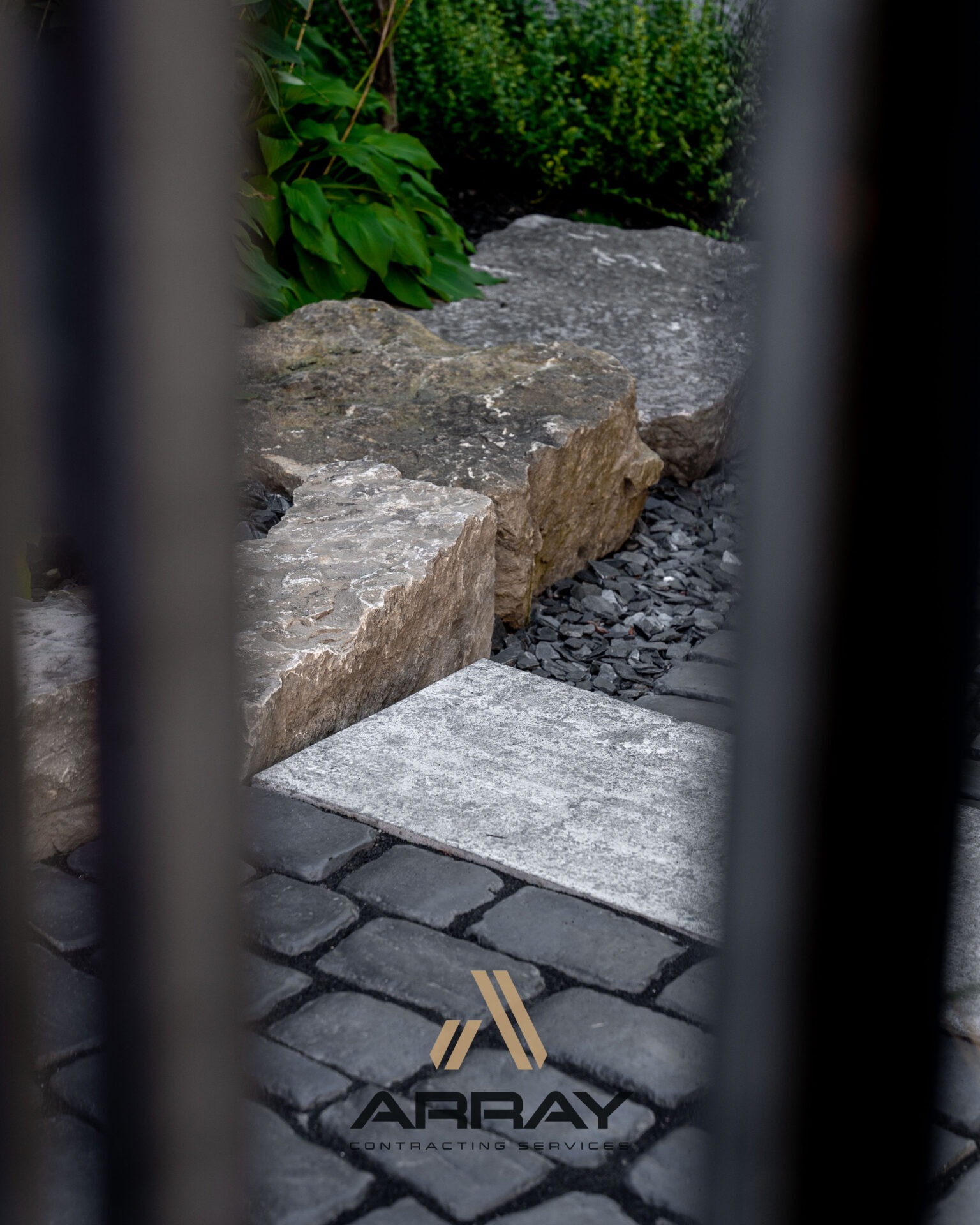 Stone landscaping with large rocks and cobblestone path, framed by black bars. Overgrown greenery in the background. Logo reads 