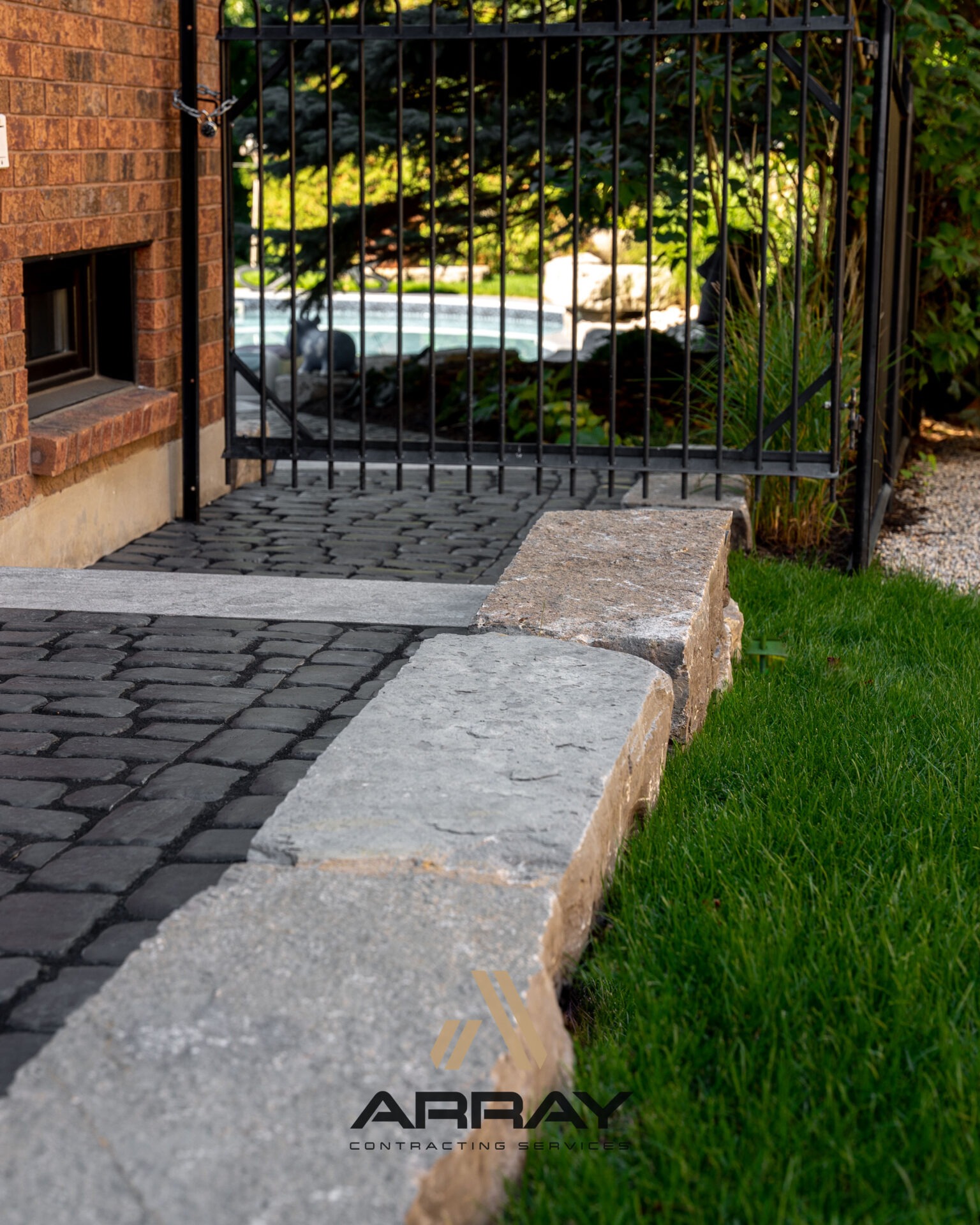 Stone pathway with grass border leads to black metal gate. Beyond, a blurred view of a pool and greenery is visible.