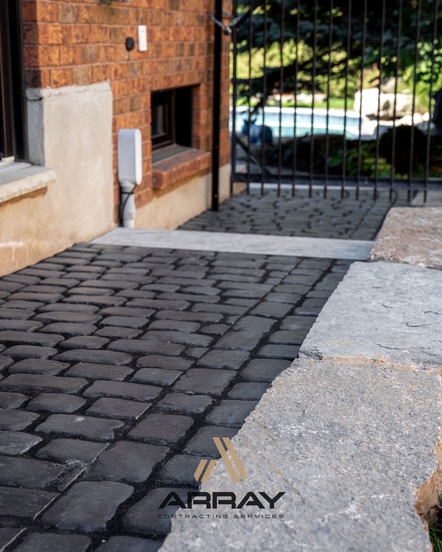 Cobblestone pathway leads to a gated area beside a brick building, featuring a small window and a distant view of greenery.