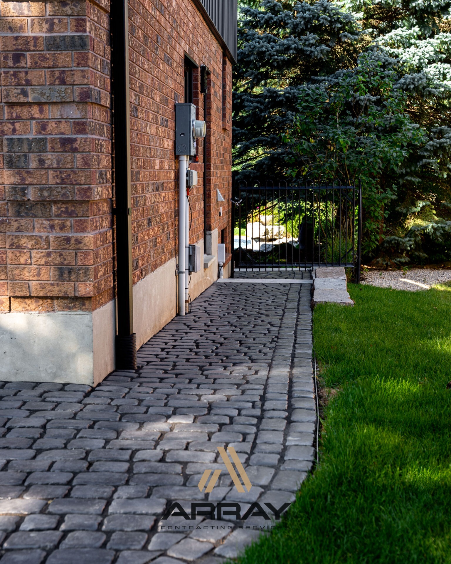 A cobblestone path runs alongside a brick house, leading to a black metal gate amidst lush greenery and vibrant grass.