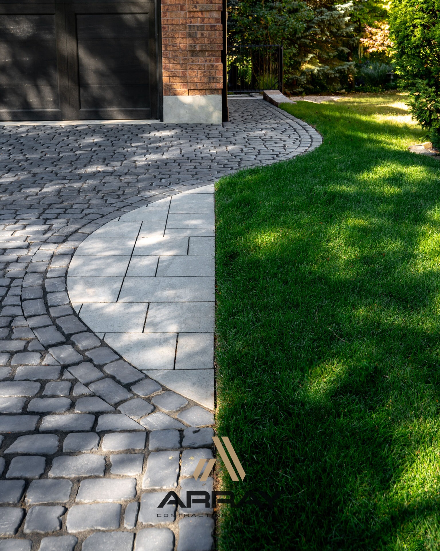 A brick driveway meets a lush, green lawn with sunlight casting shadows. The area is bordered by shrubs and trees.