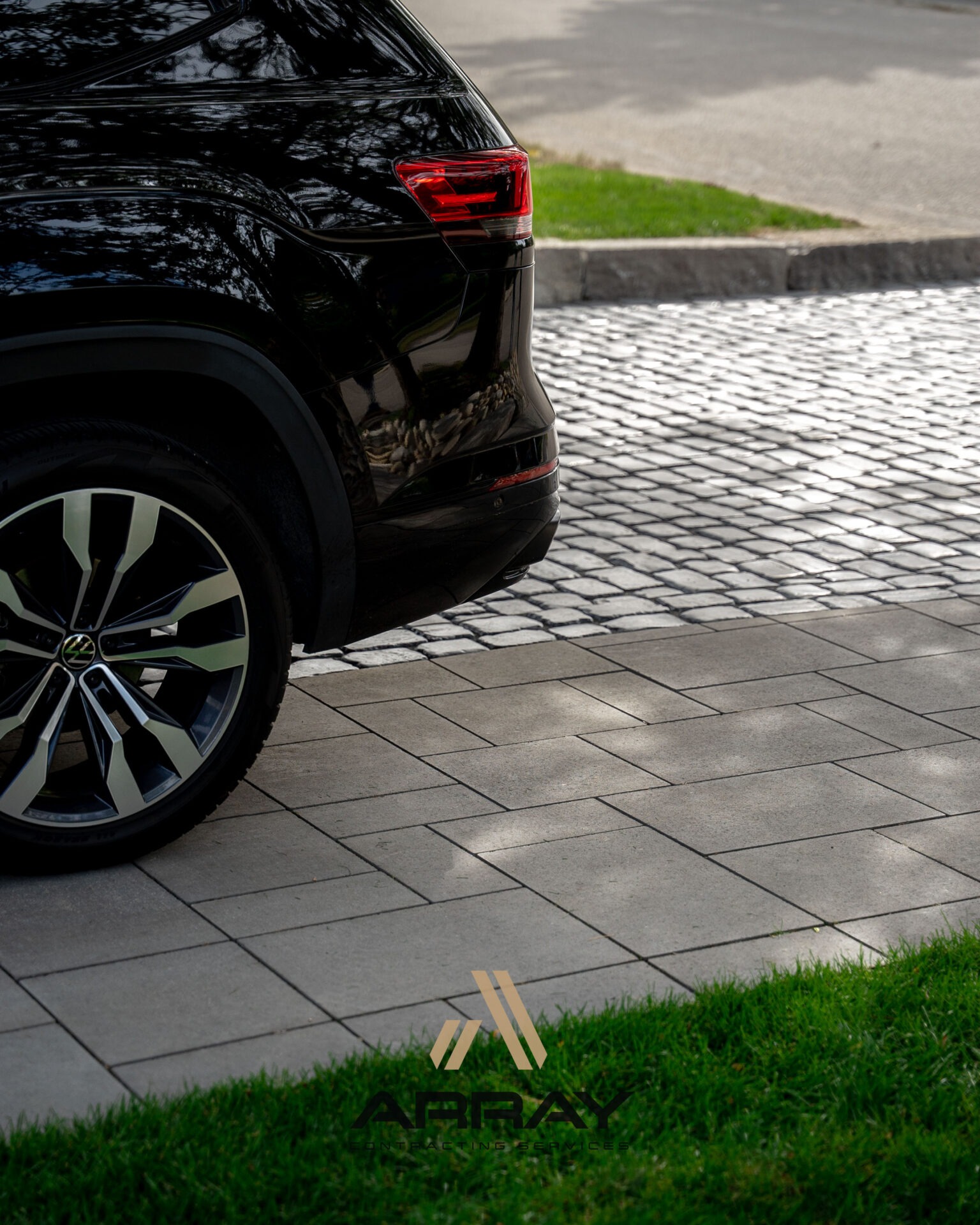 The image shows the rear section of a black car on a stone-paved driveway, adjacent to a green grass patch.