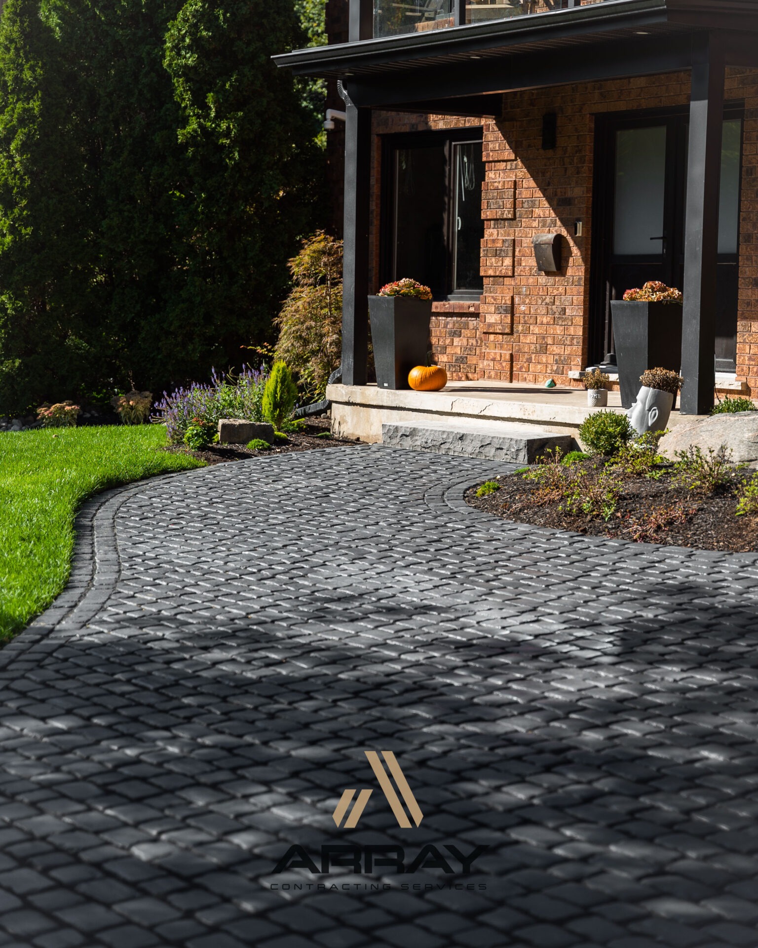 A brick house with black-trimmed windows, a stone walkway, green lawn, potted plants, and a pumpkin on the doorstep.