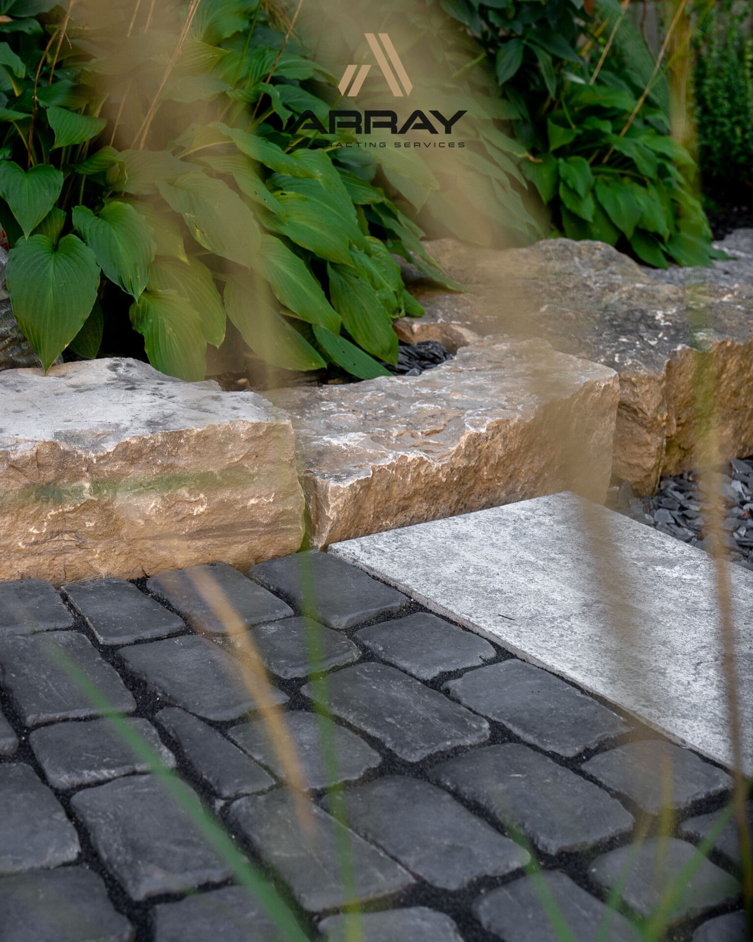 Stone path with cobblestones, bordered by large rocks and lush green foliage. Grasses gently sway in the foreground.