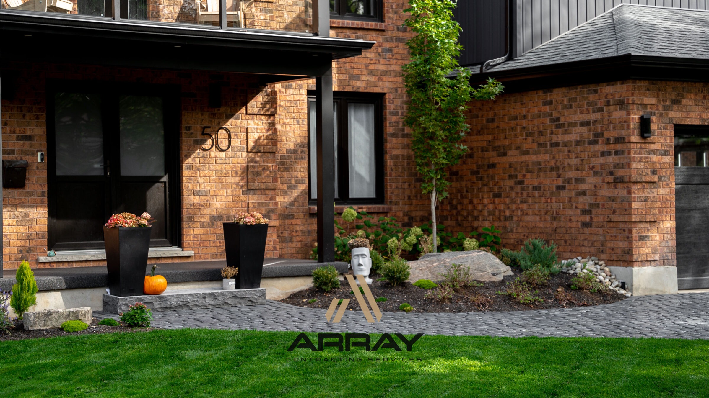 A two-story brick house with modern landscaping features a pumpkin, two tall planters, and a decorative head planter near the entrance.