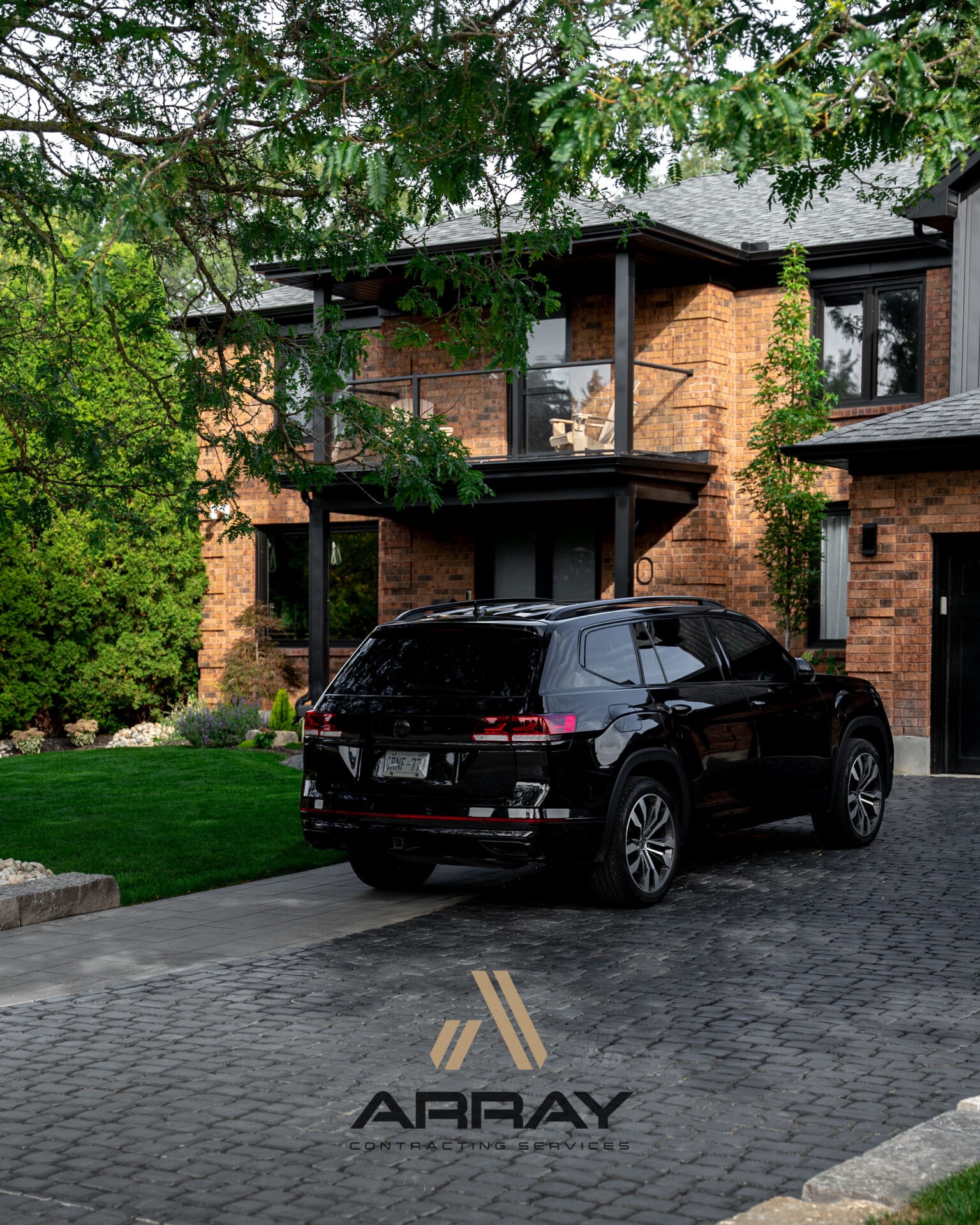 A sleek black vehicle parked in front of a modern brick house, surrounded by greenery and cobblestone paving under a leafy tree.