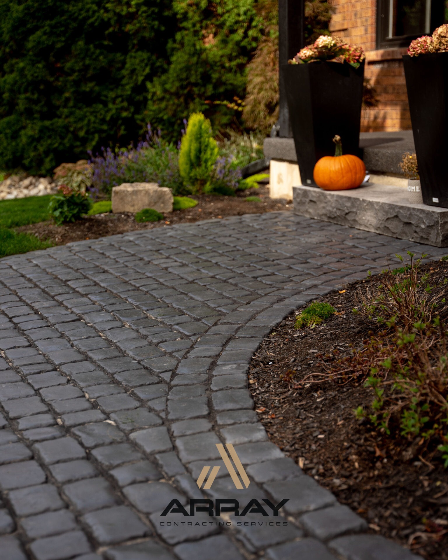 Cobblestone pathway leads to a brick porch with a pumpkin and potted plants. Lush greenery surrounds the scene, creating a welcoming atmosphere.