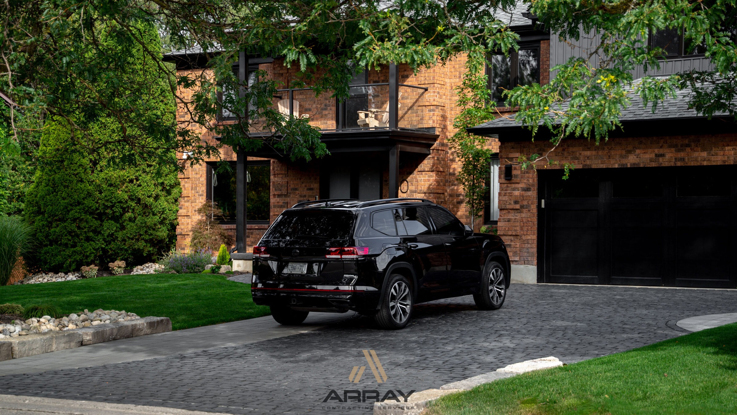 A black SUV is parked on a paved driveway in front of a modern brick house surrounded by lush greenery.