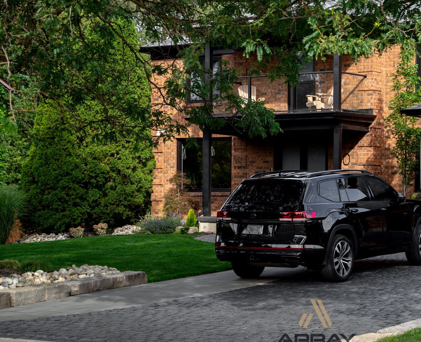 Brick house with black garage doors, surrounded by green trees. A black SUV is parked on the cobblestone driveway. No people visible.
