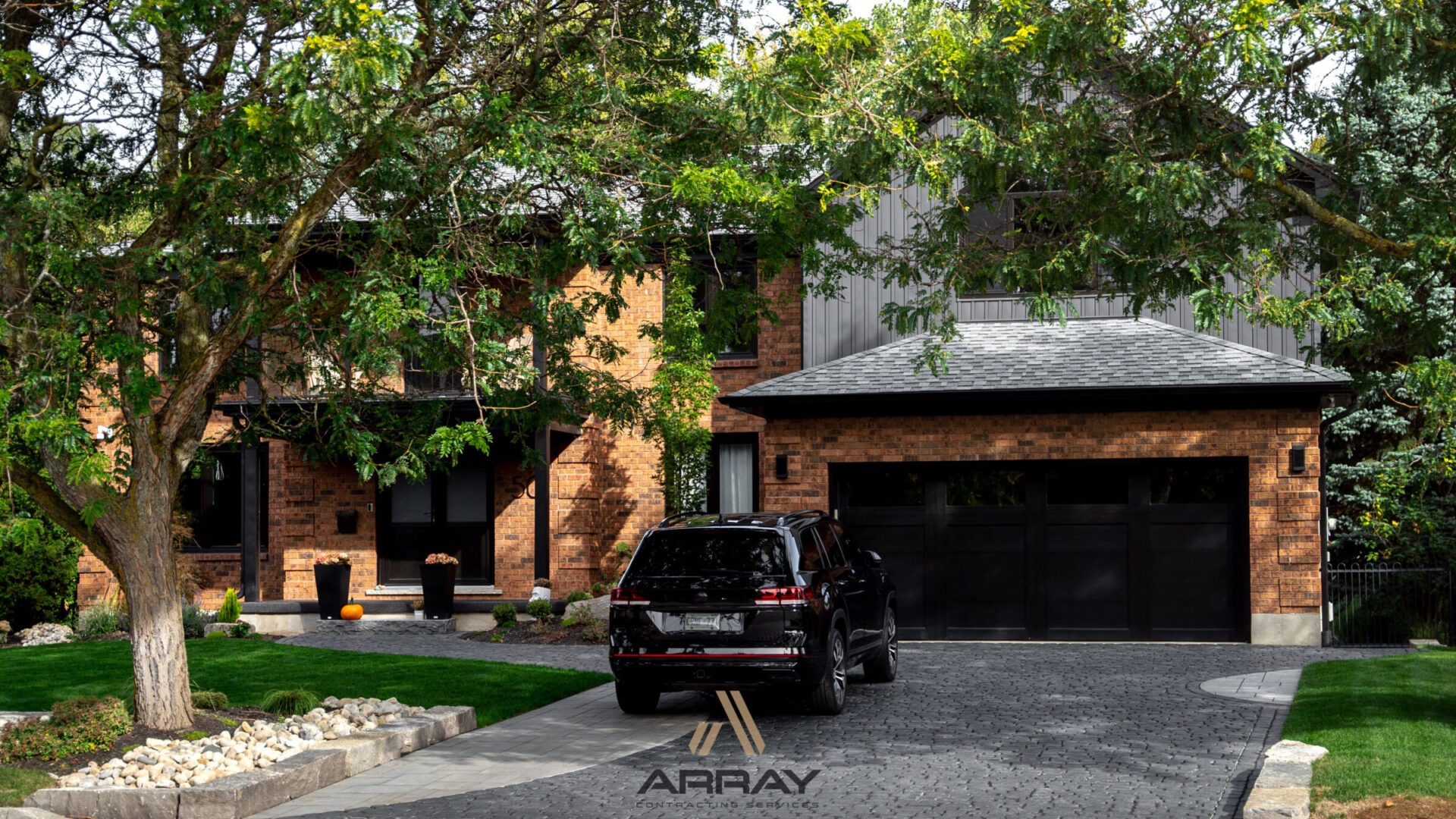 A modern brick house with a black SUV parked on the driveway, surrounded by greenery and decorative landscaping, under bright daylight.