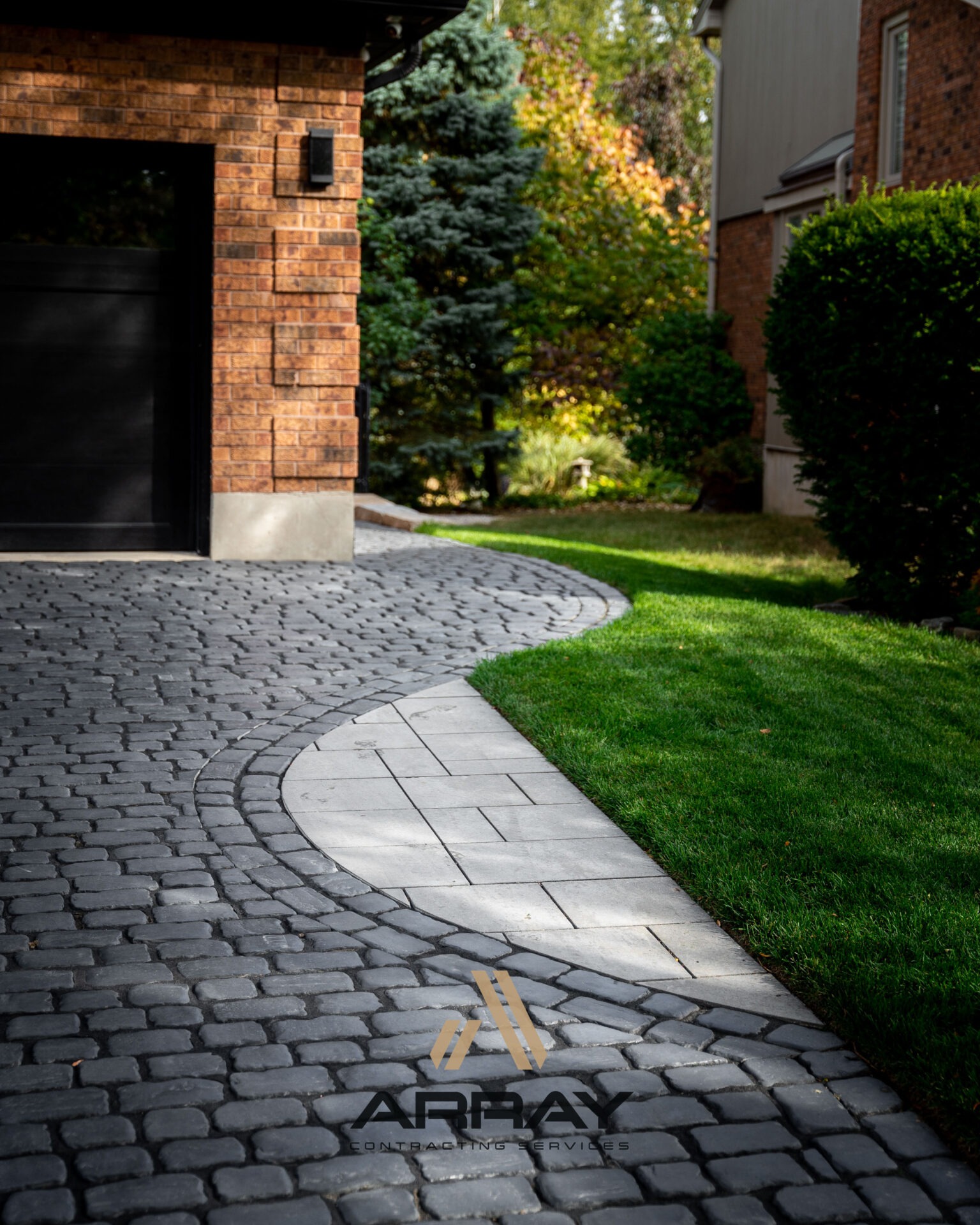 Brick house with stone pathway leading to garage entrance. Lush green lawn and bushes surround the area, suggesting a suburban residential setting.