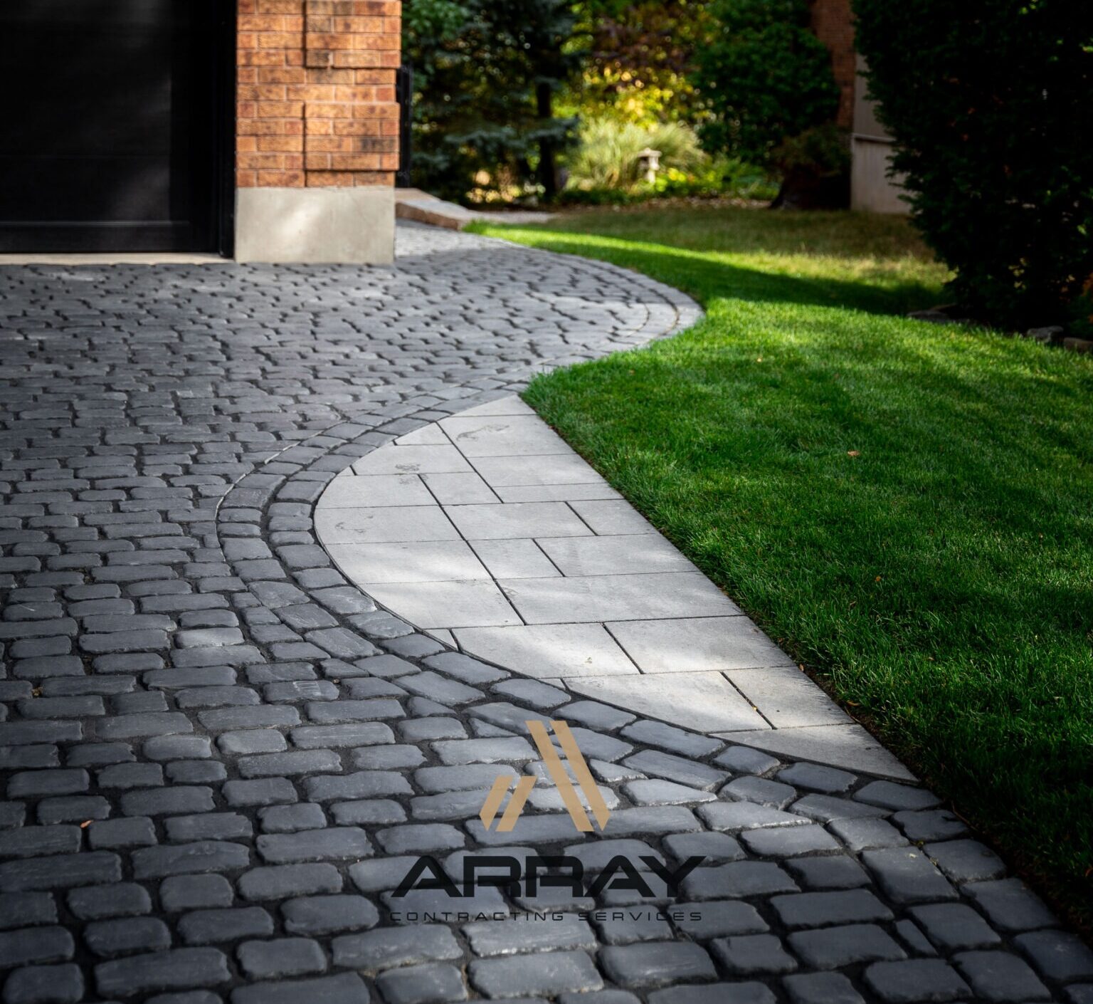 Cobblestone driveway with curved design adjacent to a grass lawn. Brick building edge visible. Logo: Array Contracting Services. Sunny, well-maintained landscape.