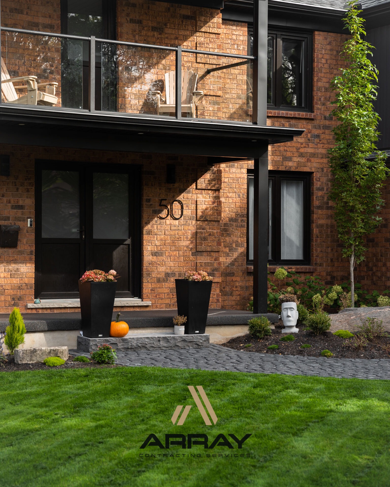 A modern brick house with a patio, decorative plants, and a pumpkin. A sculpture head placed alongside a well-maintained garden path.