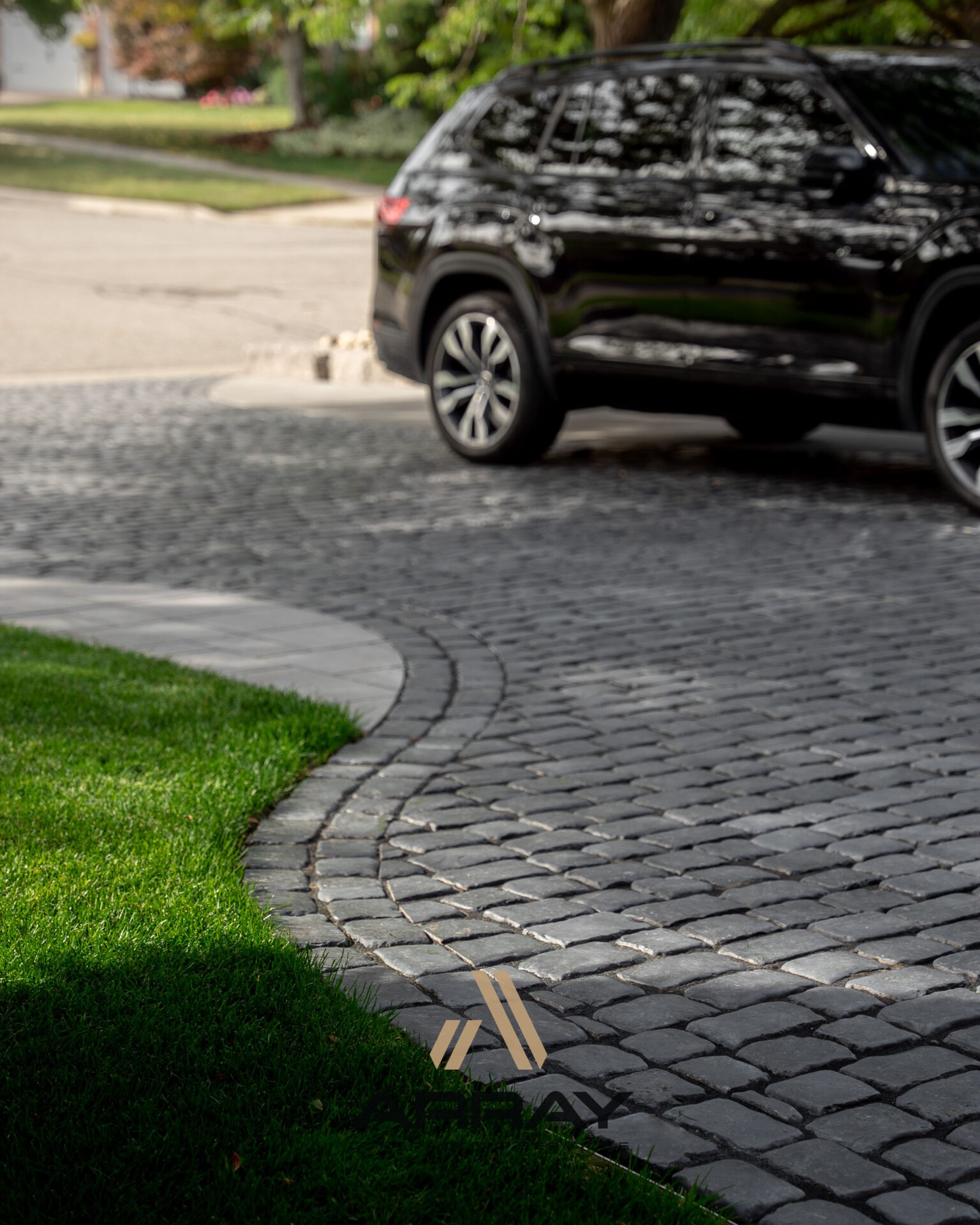 A black SUV is parked on a cobblestone driveway bordered by lush green grass and trees, under a sunny, tranquil street.