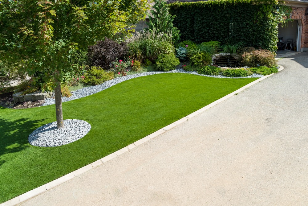 Lush green lawn with a tree, bordered by gravel. Surrounded by vibrant plants and a curved path, adjacent to a building.
