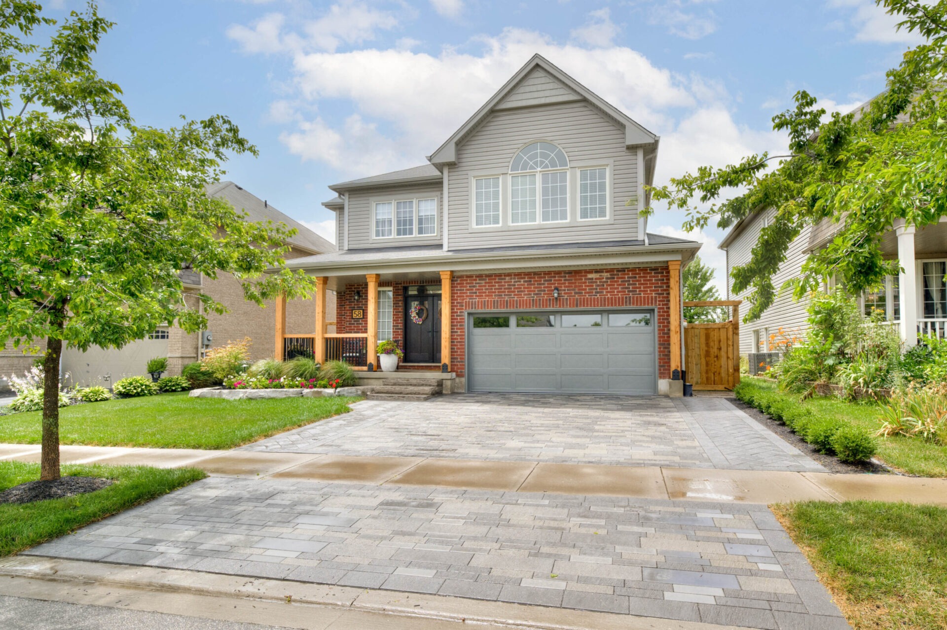 Two-story suburban house with a brick facade, gray garage, and a well-maintained garden. Clear sky and a few surrounding trees visible.