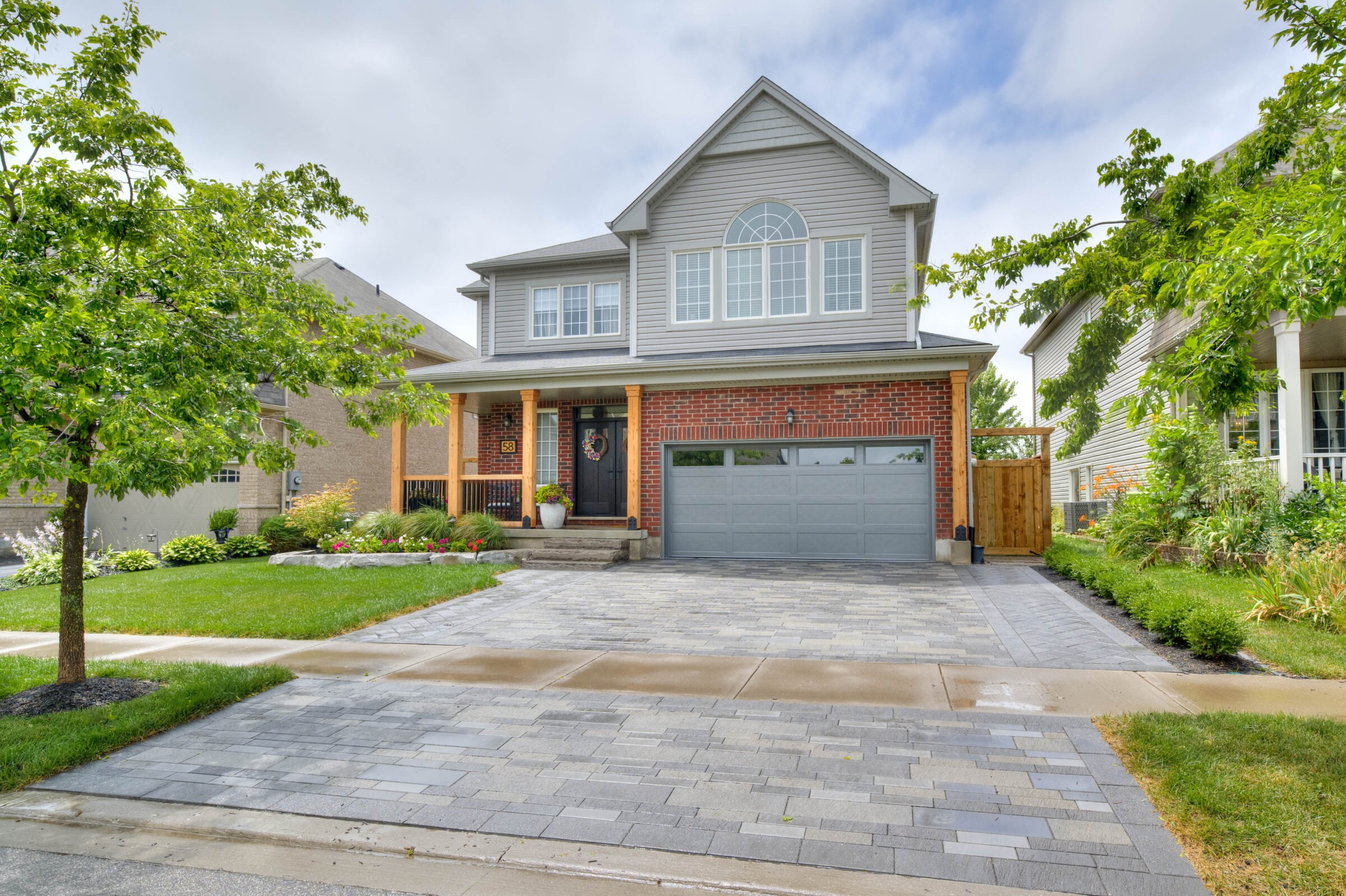 A suburban house with a red brick facade, large windows, grey garage door, lush lawn, and paved driveway, surrounded by green trees.